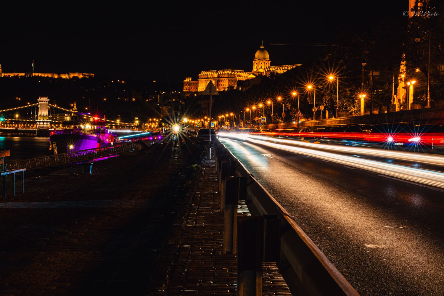 Night cityscape of Budapest with light trails on a busy road, illuminated Chain Bridge, and Buda Castle in the background.