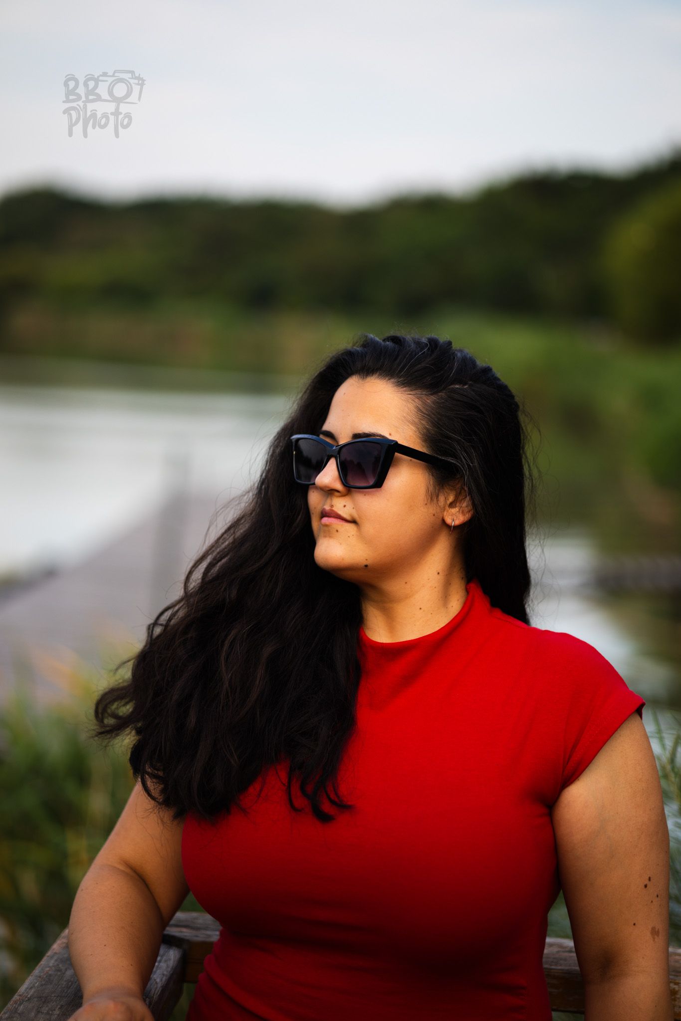 A woman with long dark hair in a red shirt and sunglasses sitting outdoors near a river.