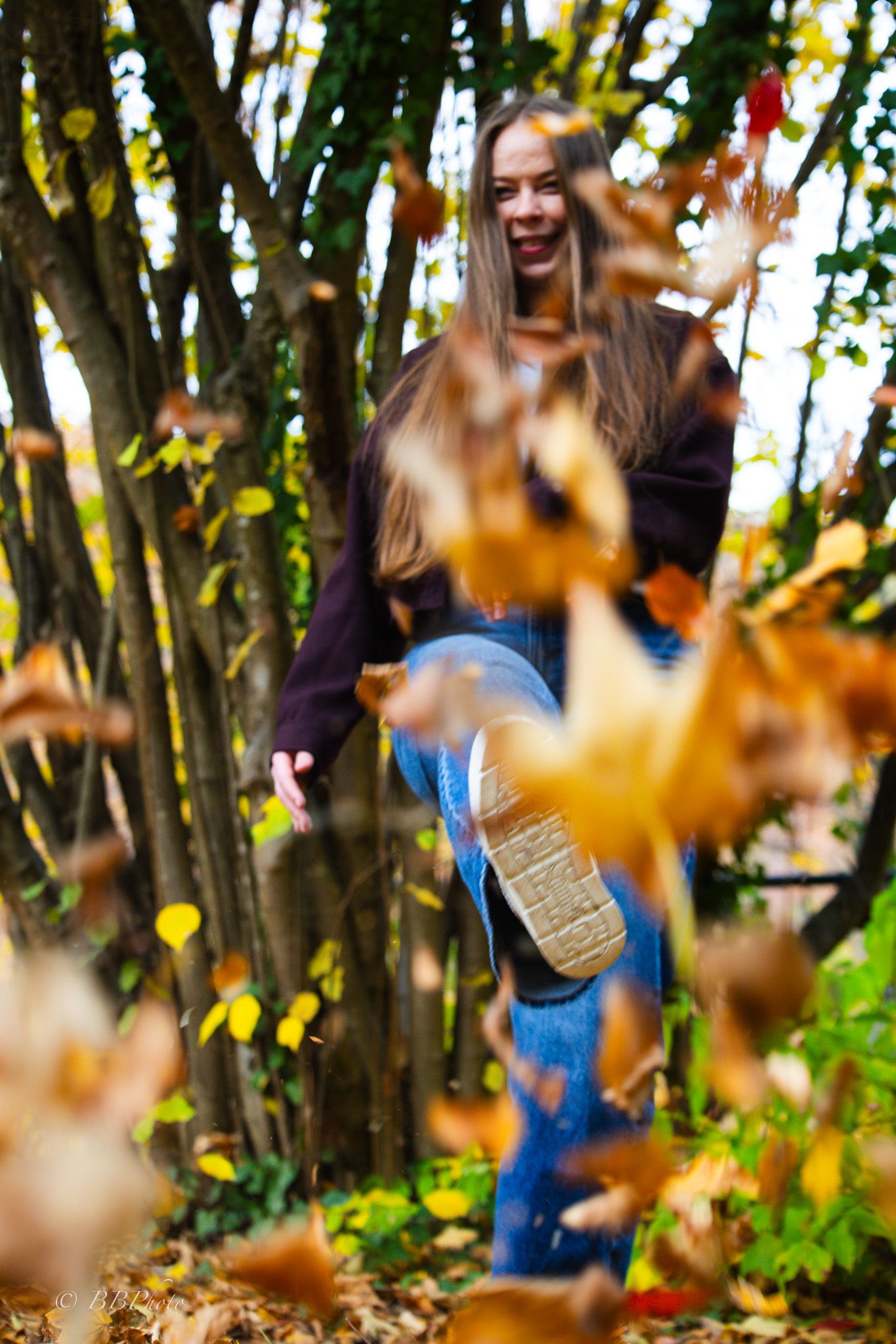 Young woman with long hair laughs while kicking up golden autumn leaves in a sunlit forest, with blurred falling leaves in the foreground.