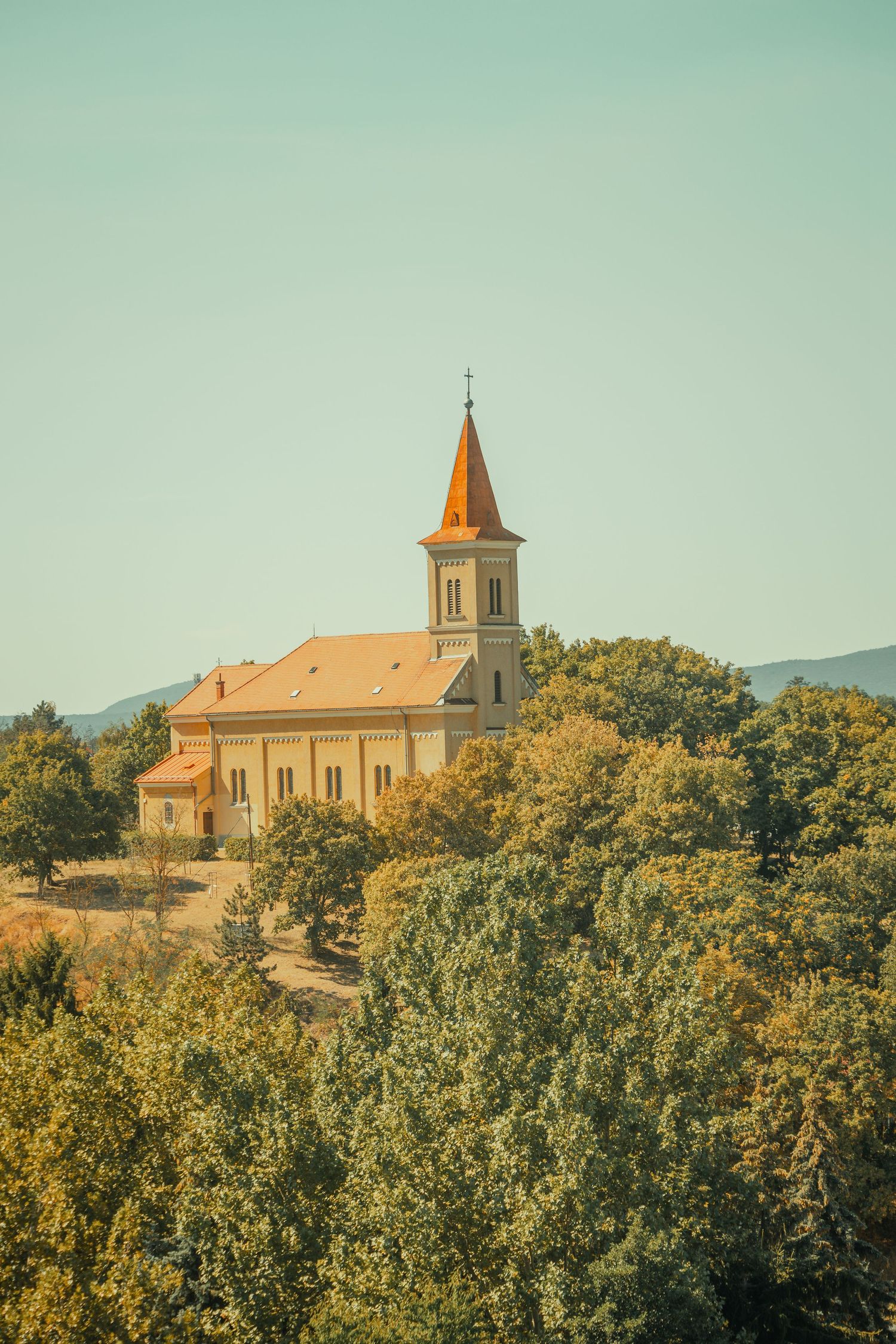 Charming rural church with orange steeple nestled among lush green trees on a hillside in Hungary.