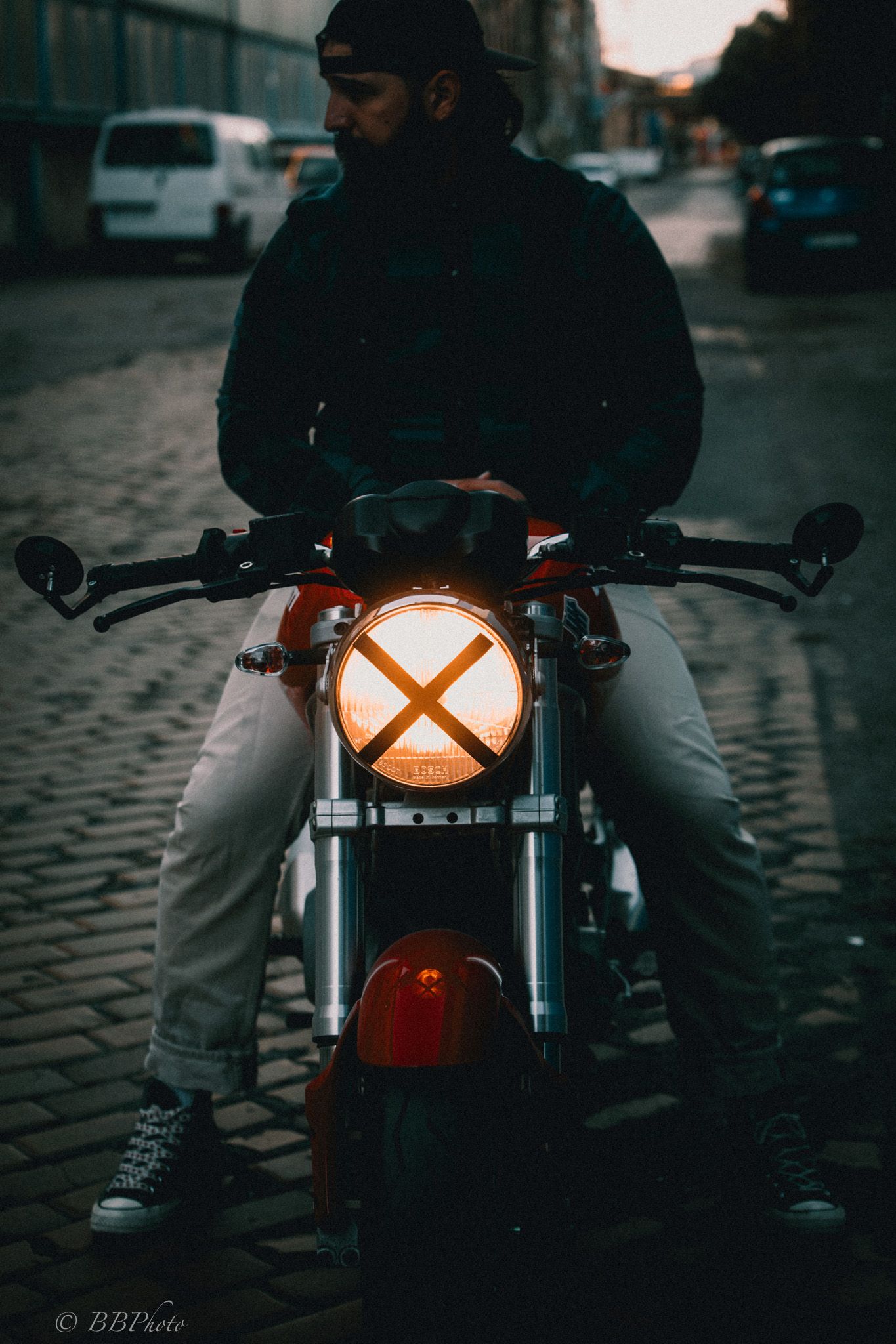 Bearded rider in a cap and dark jacket sits on a red motorcycle with a glowing X-shaped headlight on a cobblestone street at dusk.