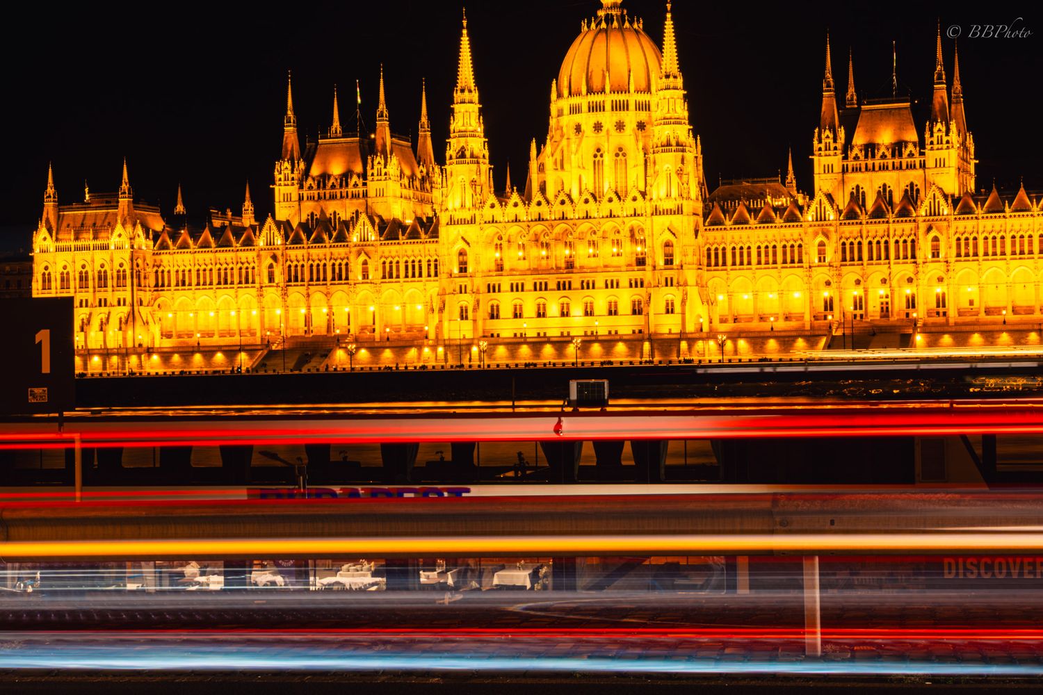 Hungarian Parliament Building illuminated in golden light at night with colorful light trails from passing boats on the Danube River.