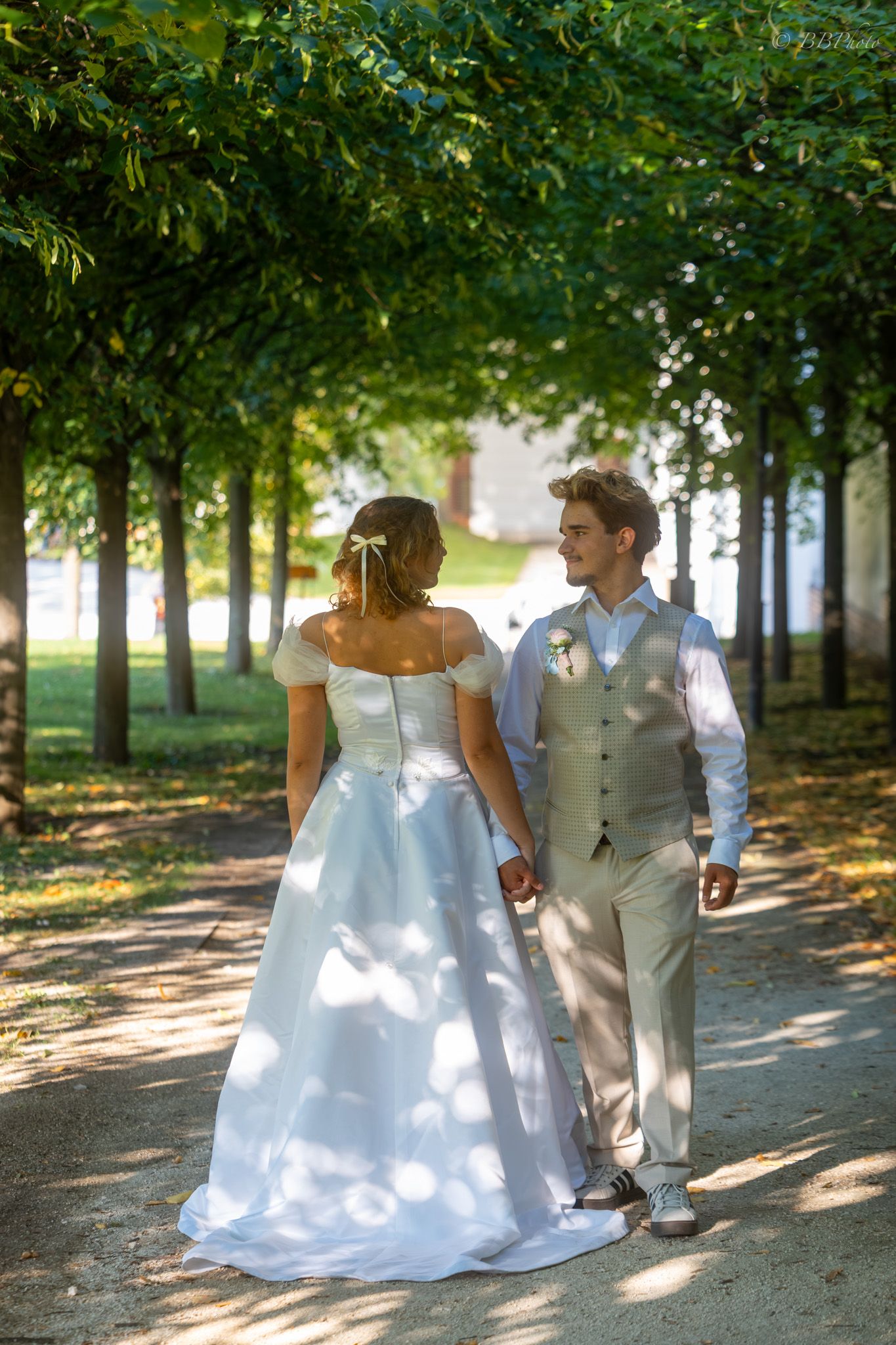 Bride and groom walk hand in hand through a sunlit tree-lined path on their wedding day.