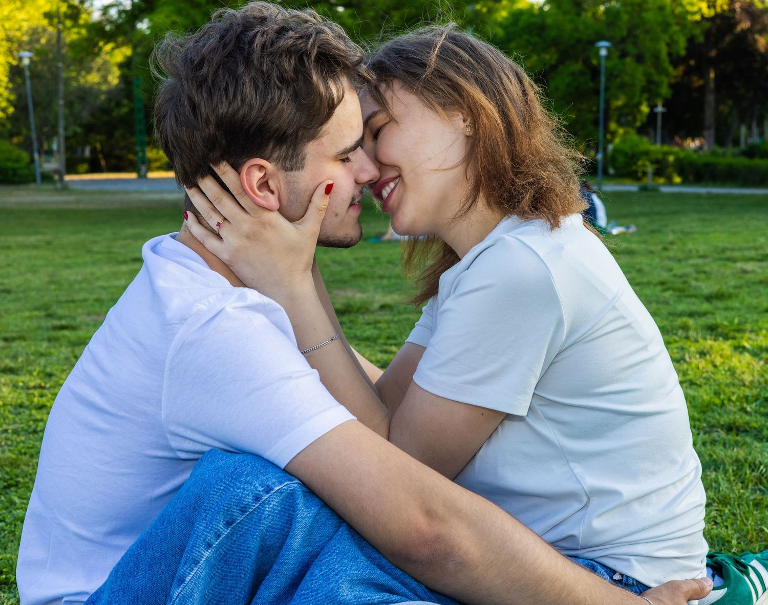 Young couple sharing a romantic moment on green grass in a sunny park, leaning in for a kiss.