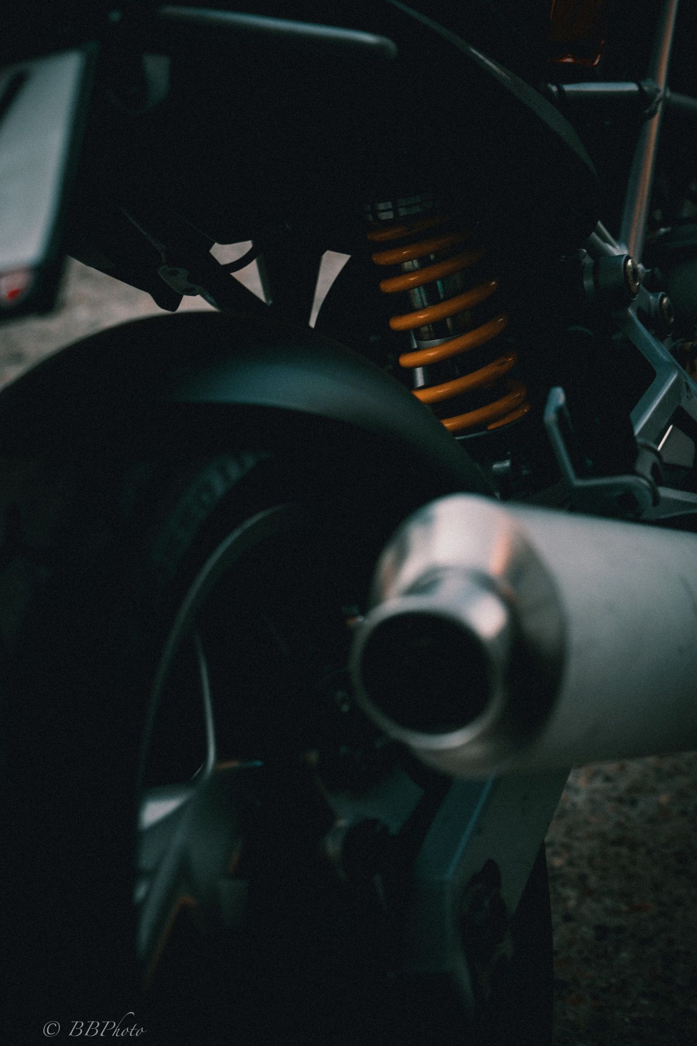 Close-up of a motorcycle's silver exhaust muffler, orange coilover rear shock absorber and rear wheel in moody low-key lighting.