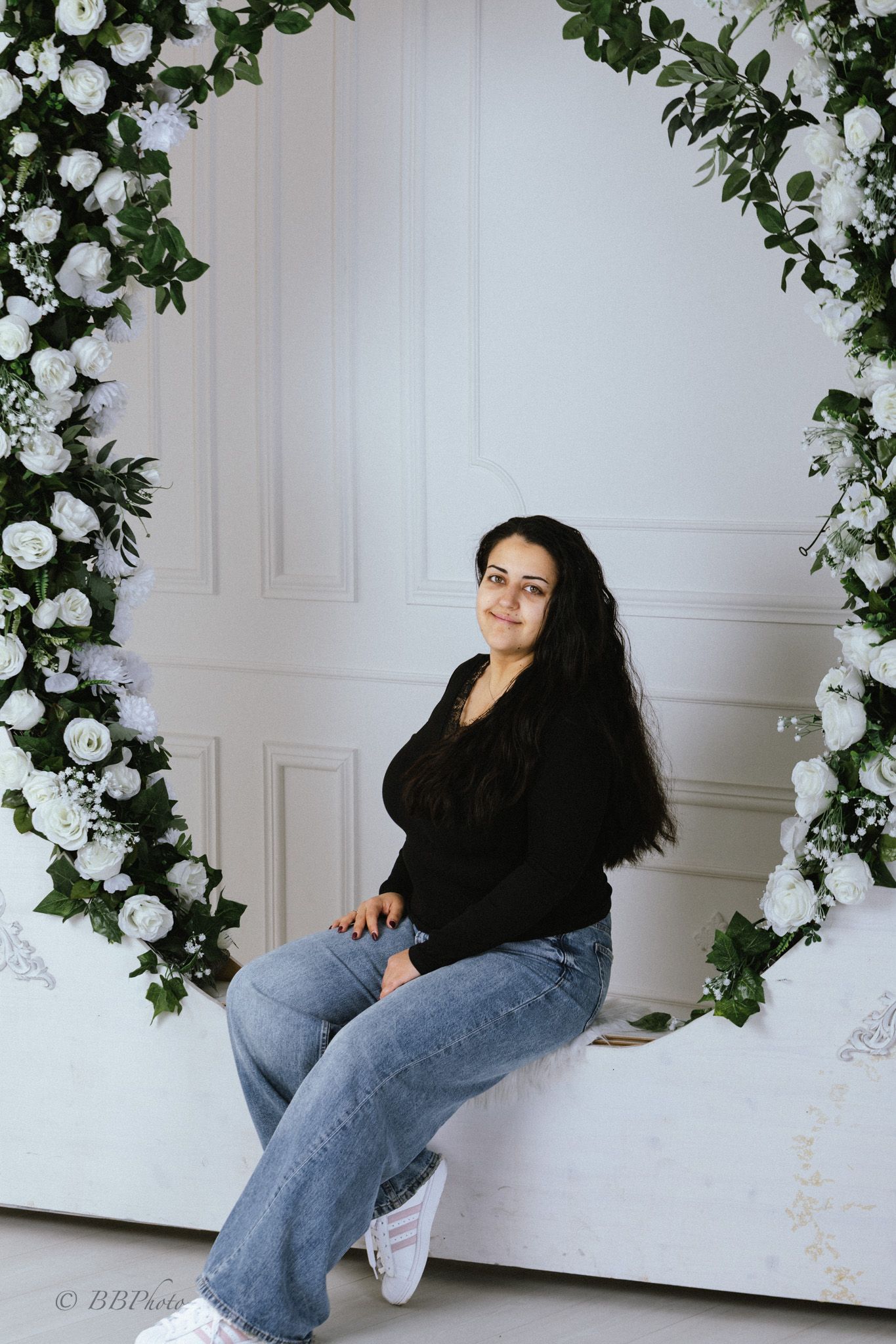 Woman in a black top and jeans sits on a white bench beneath an arch of white roses and greenery in a bright studio.