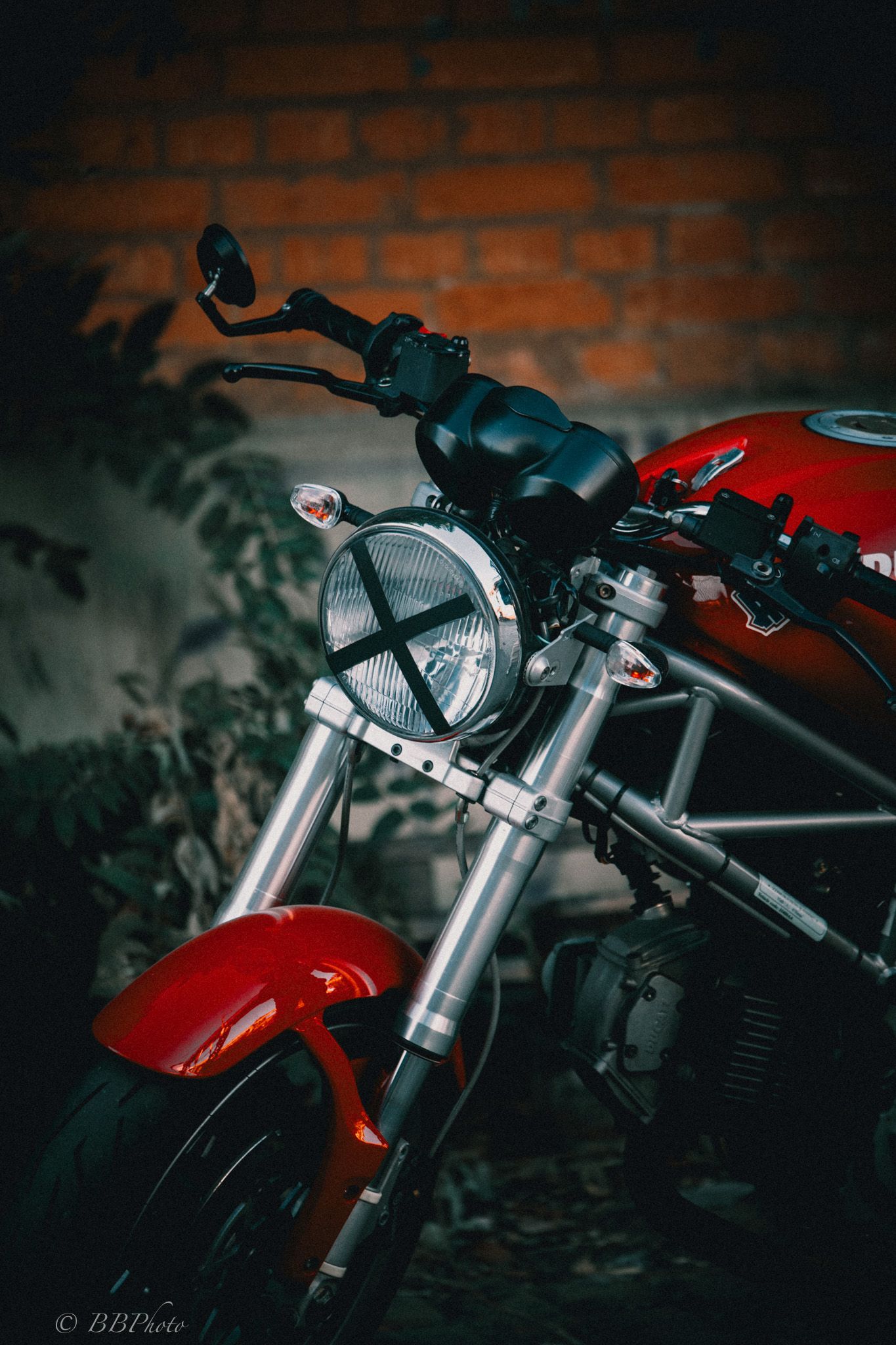 Red naked motorcycle with round headlight and X-taped lens parked in front of a rustic brick wall.