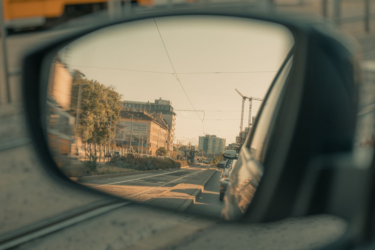 A car side mirror reflecting a city street with buildings, trees, and power lines on a sunny day.