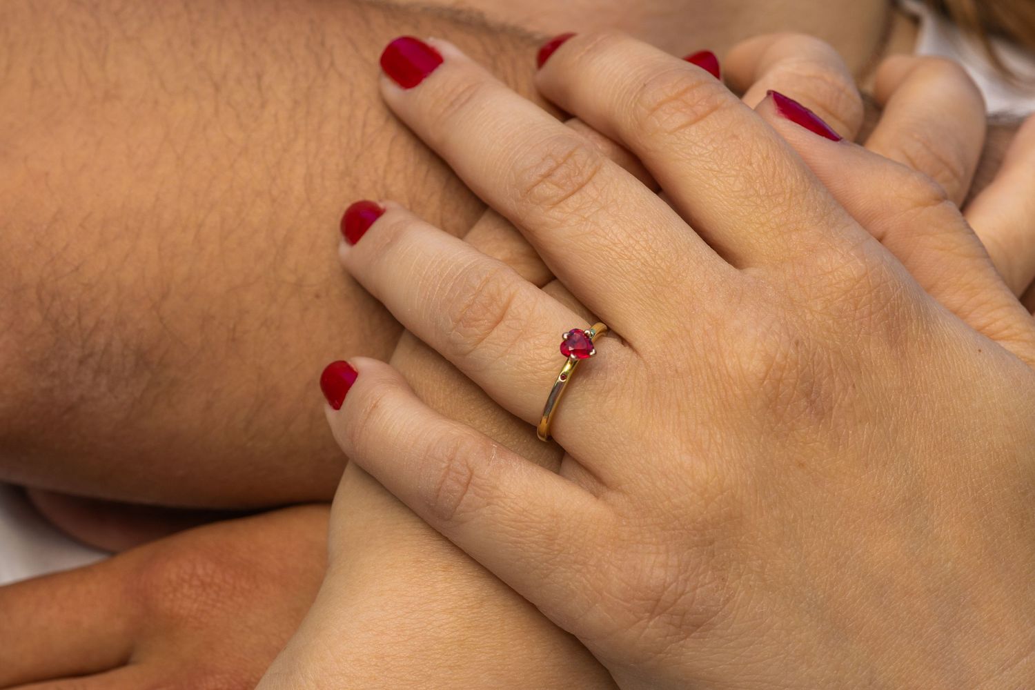 Close-up of a woman's hand with red nails wearing a gold ring with a red gemstone, resting on another person's arm.