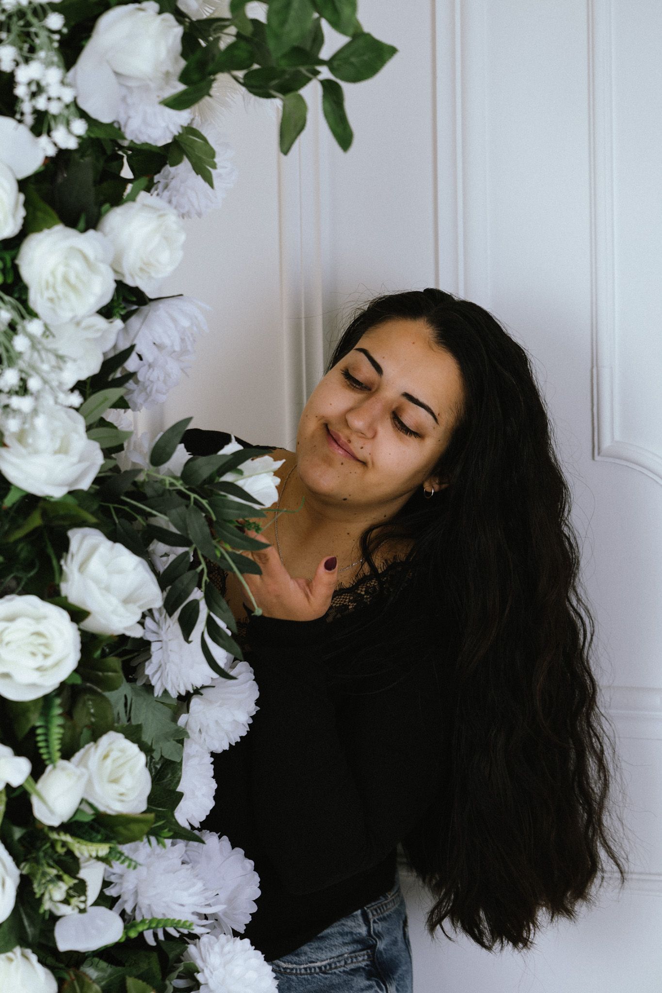 Smiling woman with long dark hair and closed eyes leans against a white paneled wall beside a lush white rose and green foliage arrangement.