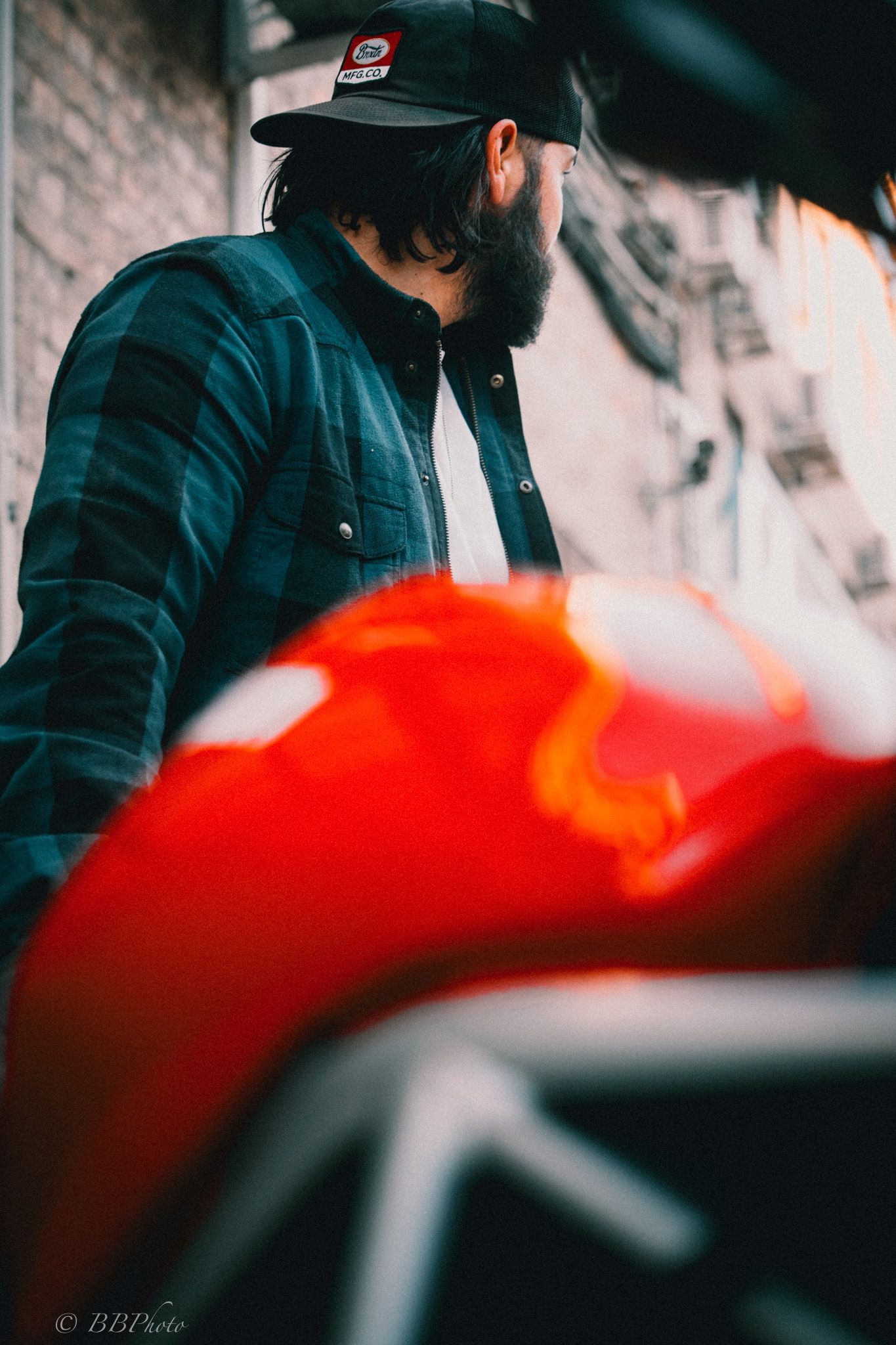 Bearded man in a black cap and green plaid flannel shirt stands beside a glossy red motorcycle fuel tank near a brick building.