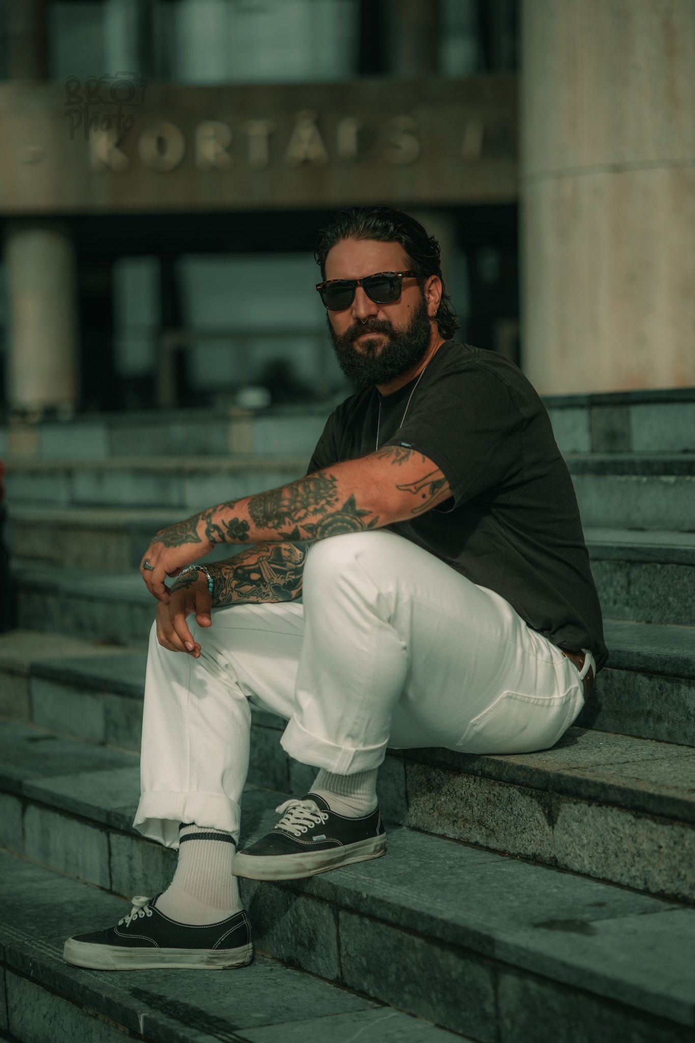 Tattooed bearded man in sunglasses, black tee, and white pants sitting on stone steps outside a building.