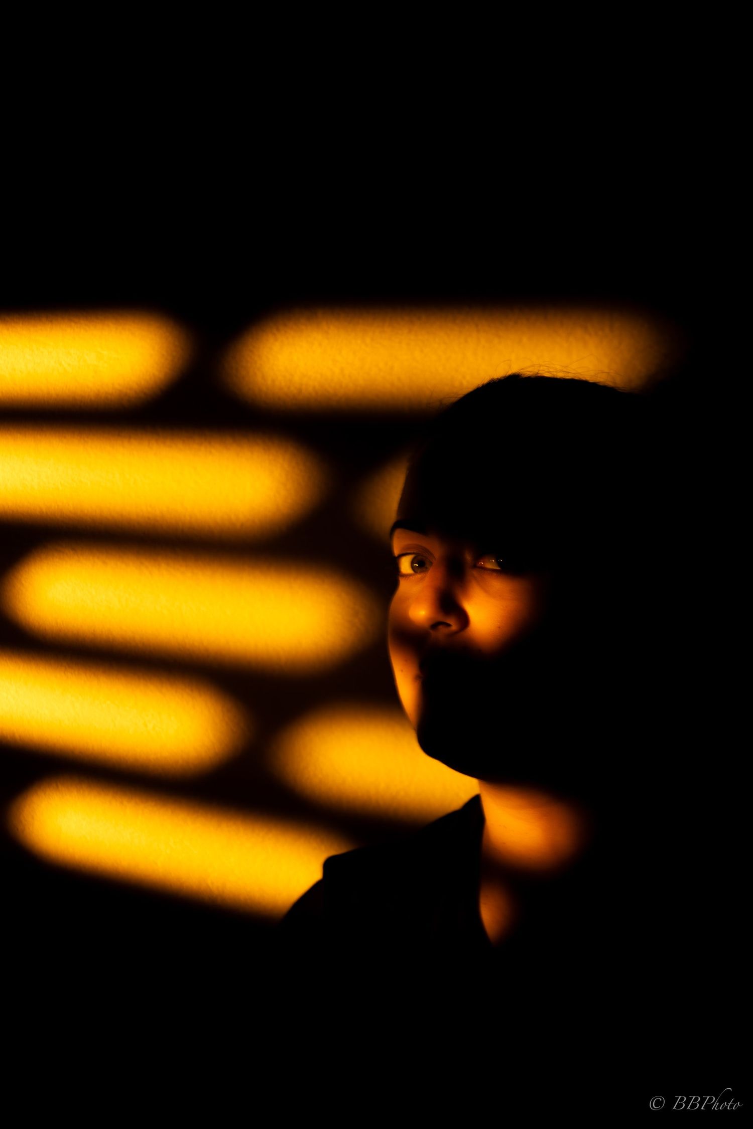 Dramatic portrait of a person silhouetted against golden light stripes from blinds on a dark wall.