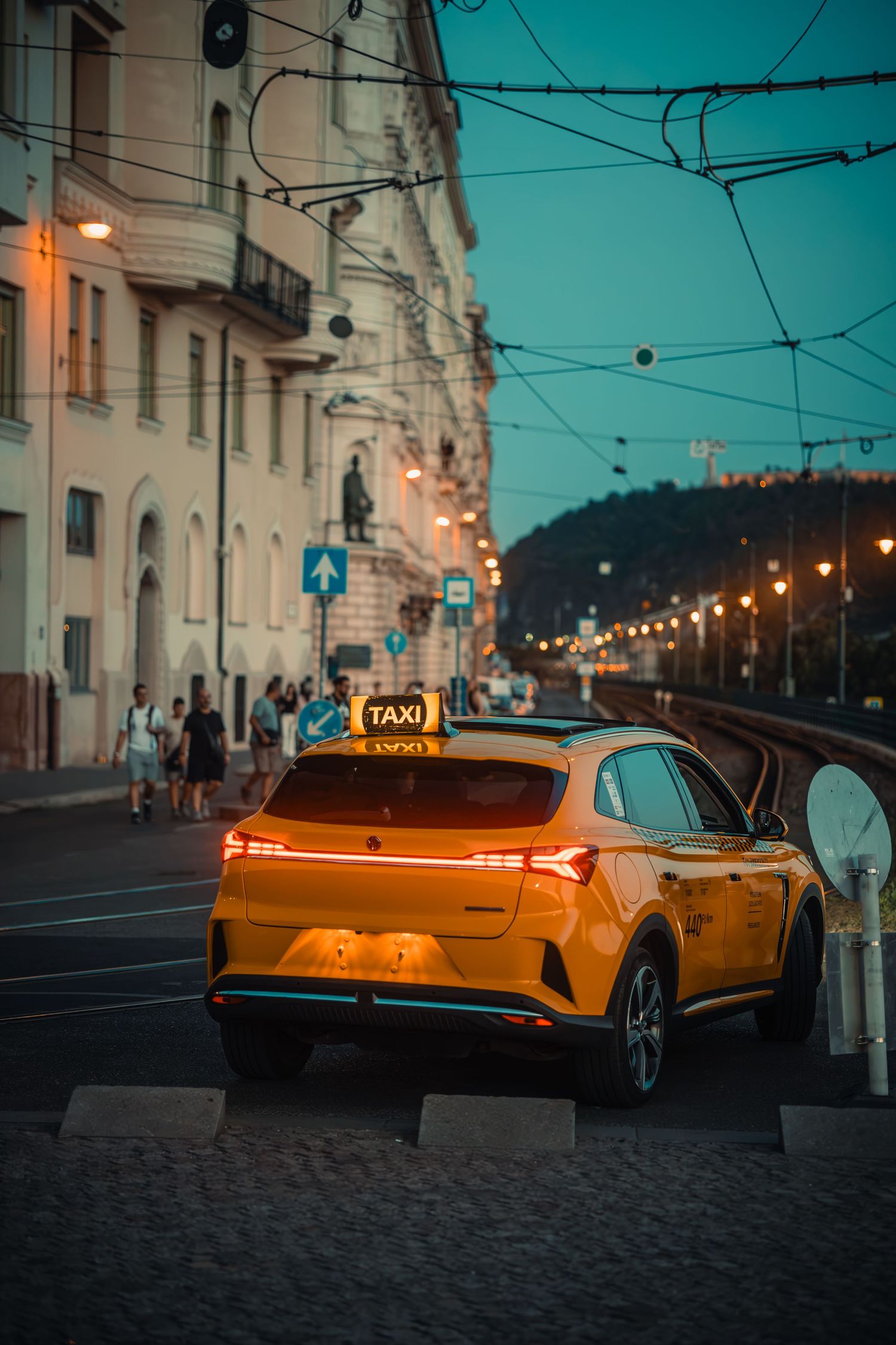 Yellow taxi cab parked on a cobblestone city street at dusk with glowing streetlights and historic buildings in background.