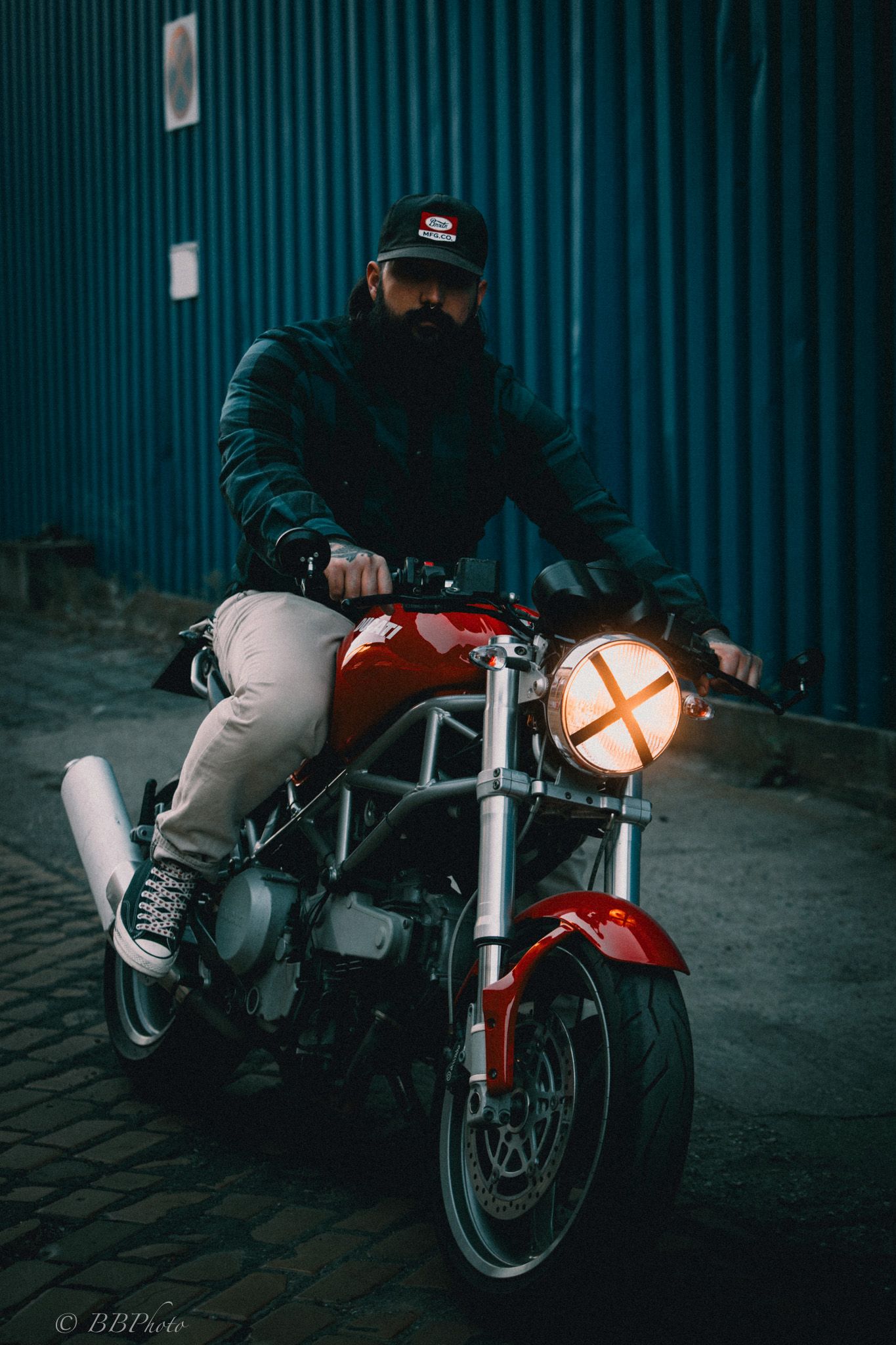 Bearded man in a black cap and leather jacket rides a red cafe racer motorcycle with a glowing cross headlight past a blue corrugated wall.