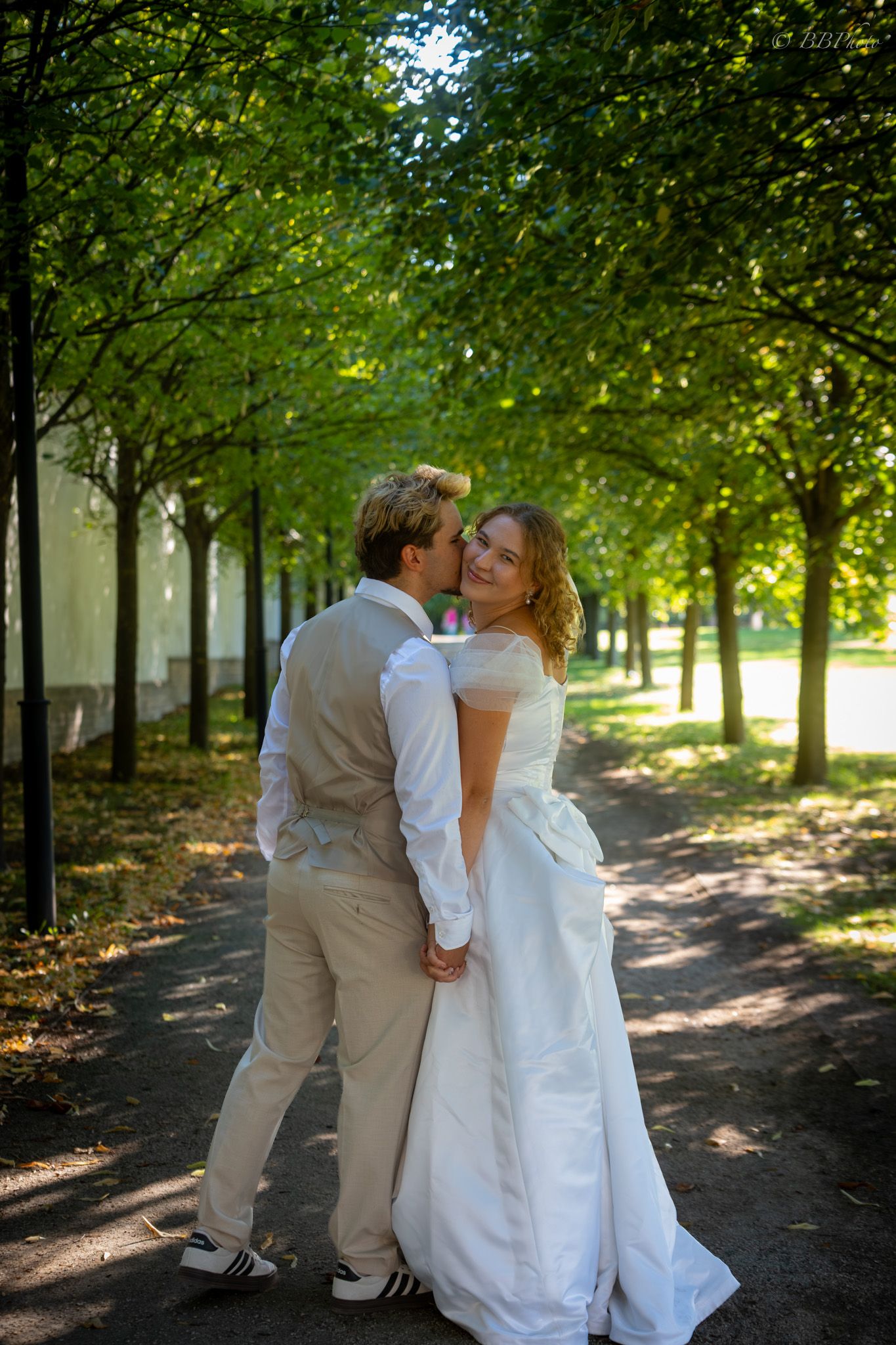 Newlywed couple embracing on a tree-lined path, bride in white gown smiling at camera on their wedding day.