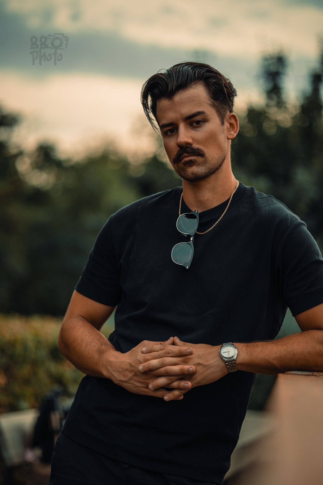 Serious man with mustache in black t-shirt with sunglasses hanging from collar at dusk outdoors.