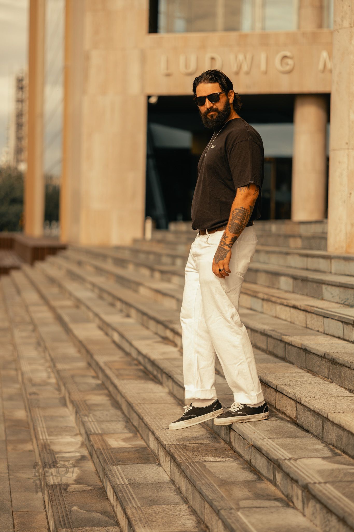 Tattooed bearded man in white pants and dark shirt posing on stone steps outside Ludwig Museum.