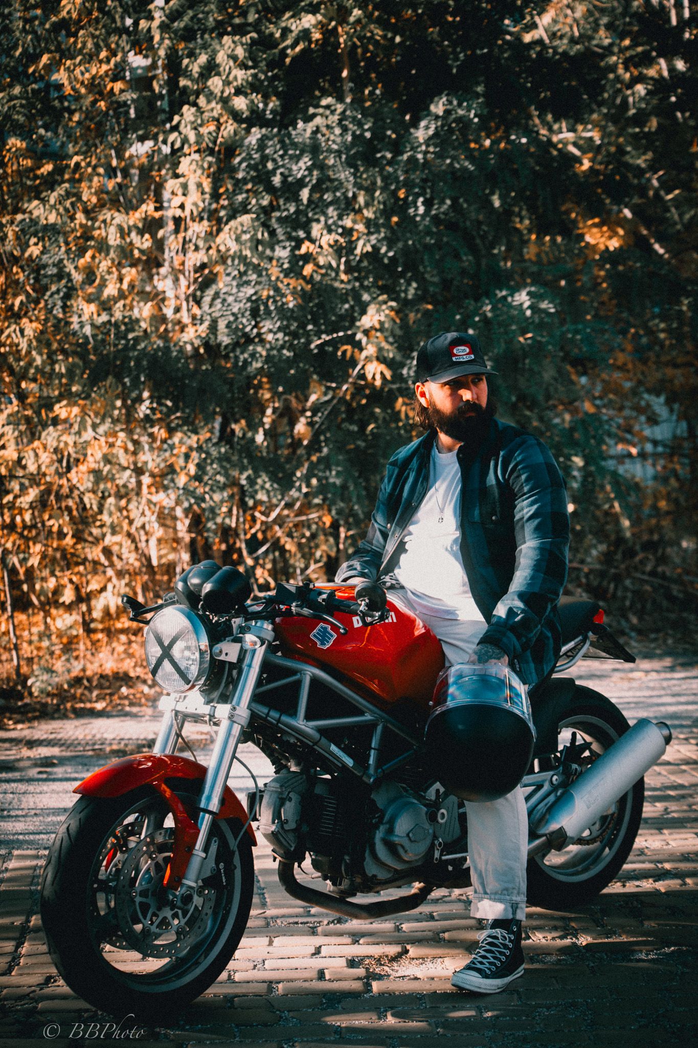 Bearded man in a black cap and leather jacket sits on a red Ducati Monster motorcycle on a brick path near autumn trees.