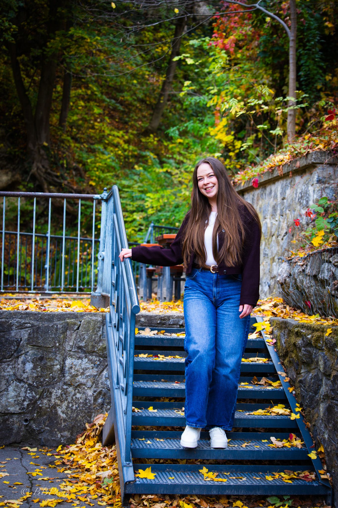 Smiling woman with long brown hair walks down blue metal steps surrounded by autumn leaves and colorful fall foliage.