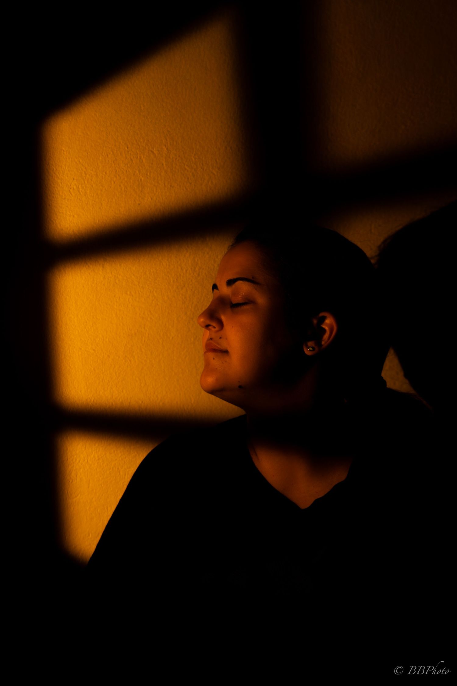 Woman bathed in warm golden window light with eyes closed, dramatic shadows creating a moody, contemplative portrait.
