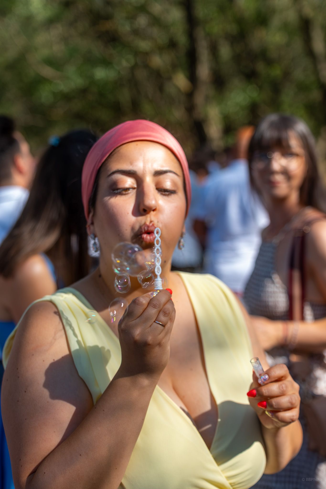 Woman in yellow dress and pink headscarf blowing soap bubbles outdoors at a sunny gathering.
