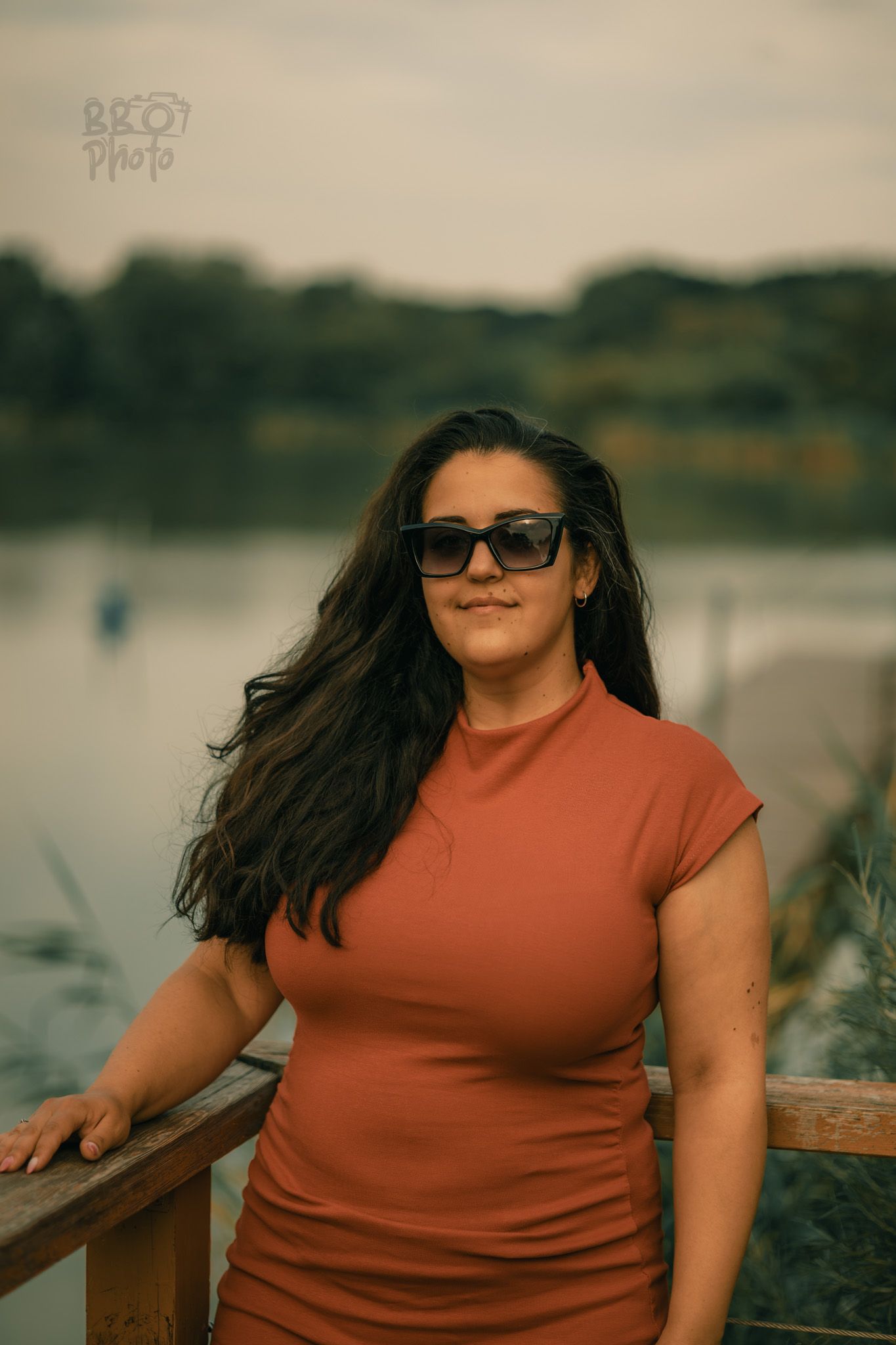 Woman with long dark hair and sunglasses in an orange dress leaning on a wooden railing by a lake.