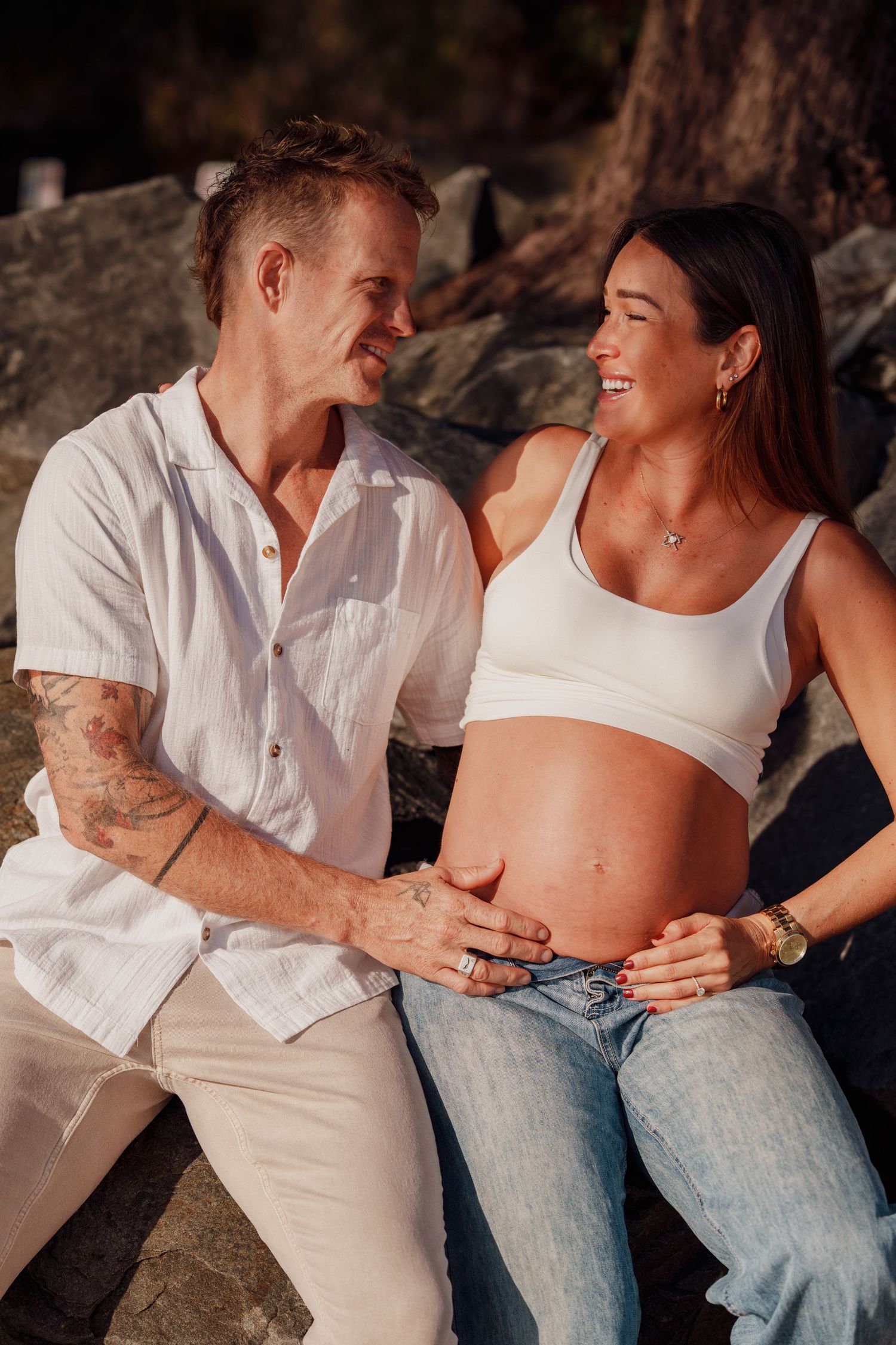 Smiling pregnant woman in white crop top and jeans with tattooed partner touching baby bump outdoors.