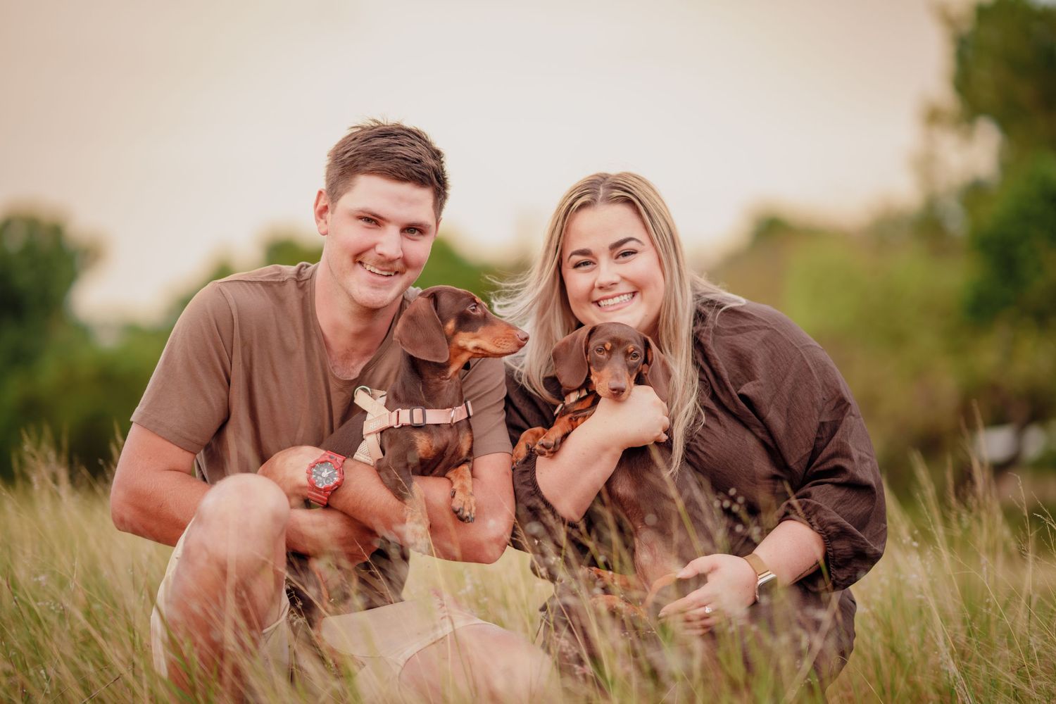 Smiling couple sitting in tall grass holding two small dachshund dogs during a golden hour outdoor photo session.