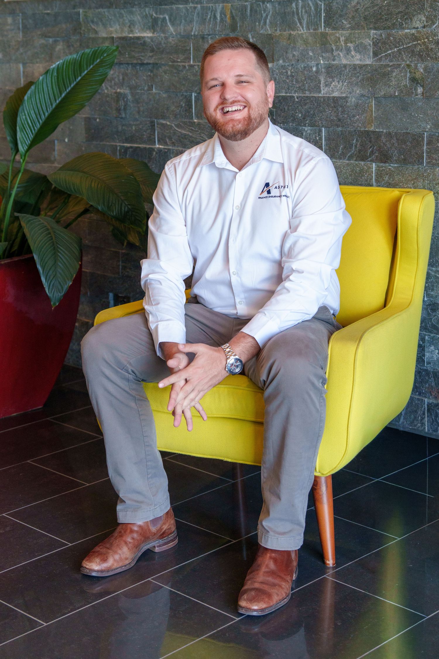 Smiling man in white shirt and khakis sits relaxed in a bright yellow chair against a stone wall with tropical plant.