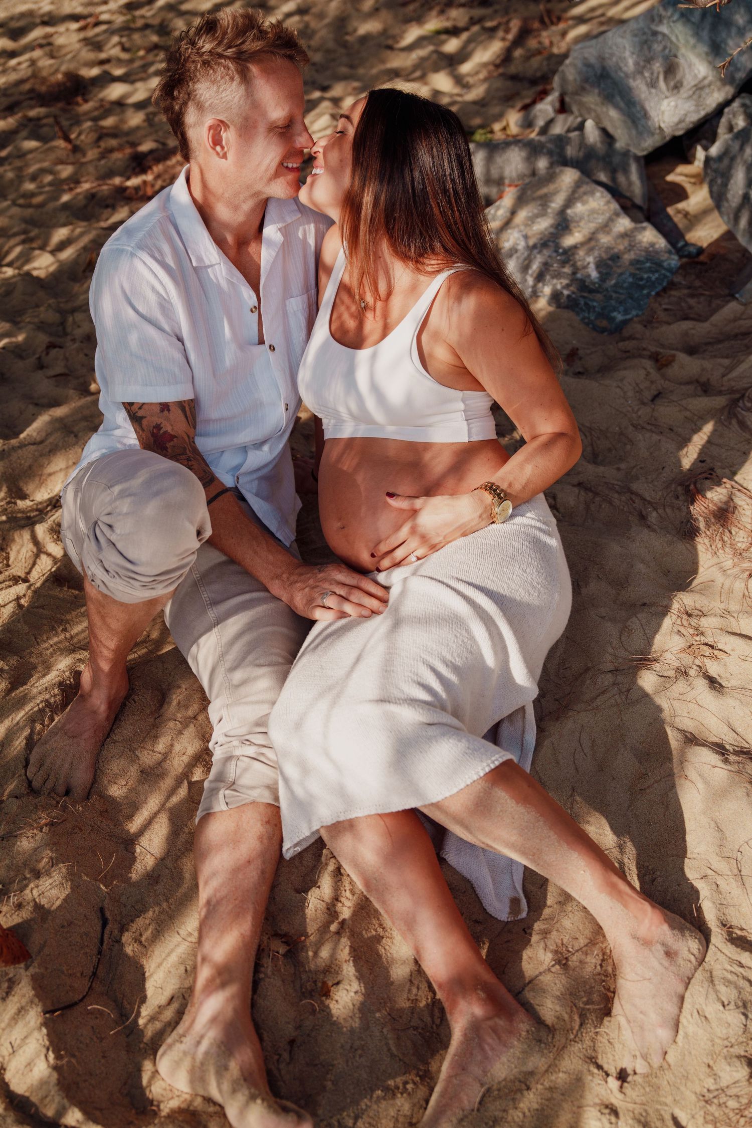 Couple in white outfits sitting on sandy beach, man embracing pregnant woman's belly in golden light.