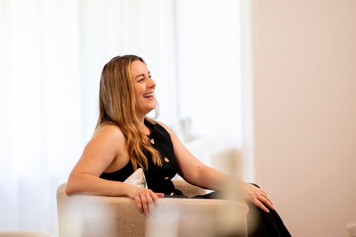 A laughing woman with long blonde hair sits relaxed on a white chair in a bright, modern room.