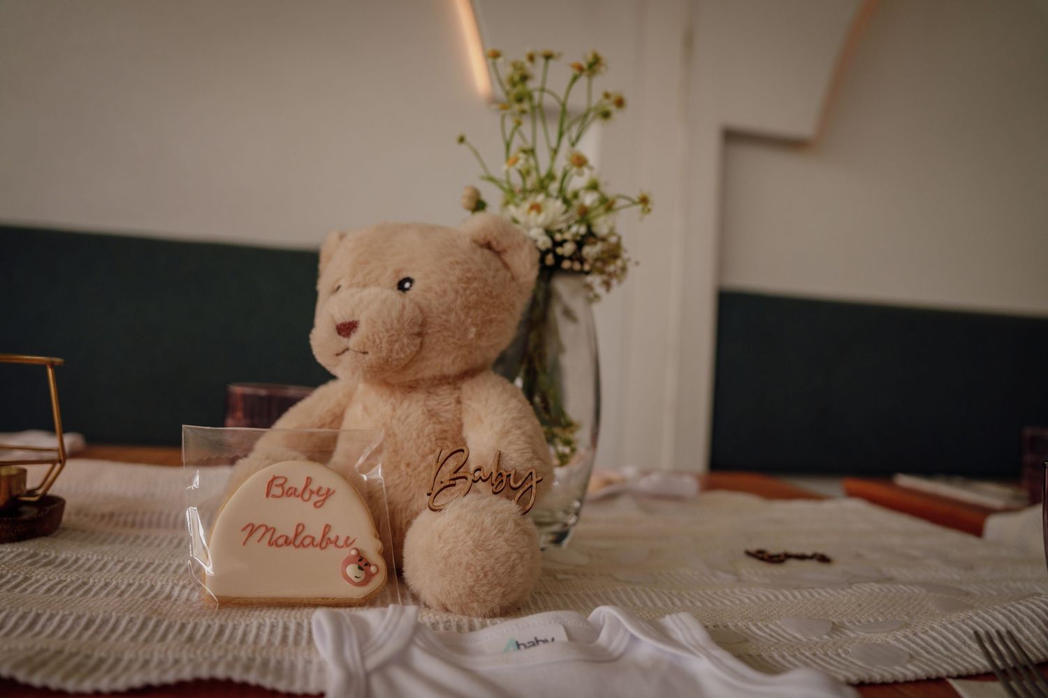 A teddy bear with embroidered name sits on a decorated table with floral arrangement at a baby shower.