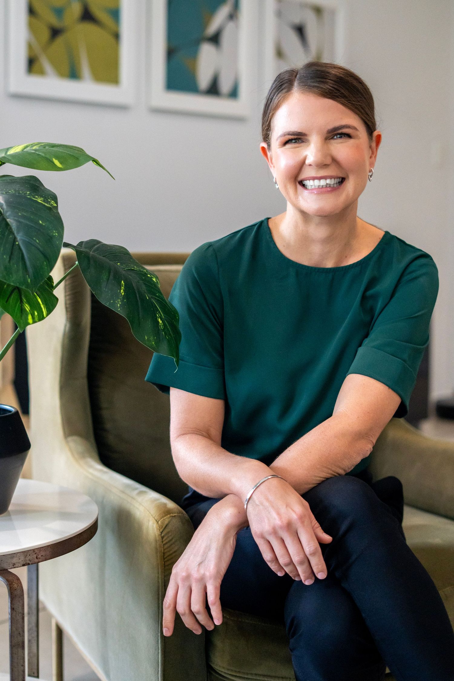Smiling woman in green top sitting relaxed in olive chair beside tropical plant in modern interior.