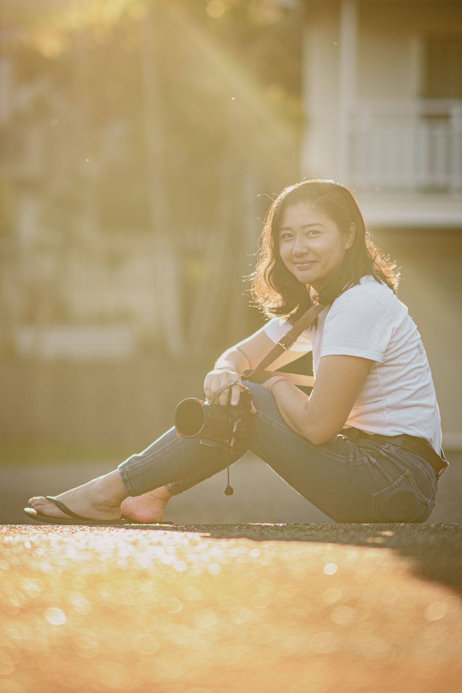 A smiling Asian woman sits outdoors holding a camera, bathed in warm golden sunlight with a blurred background.