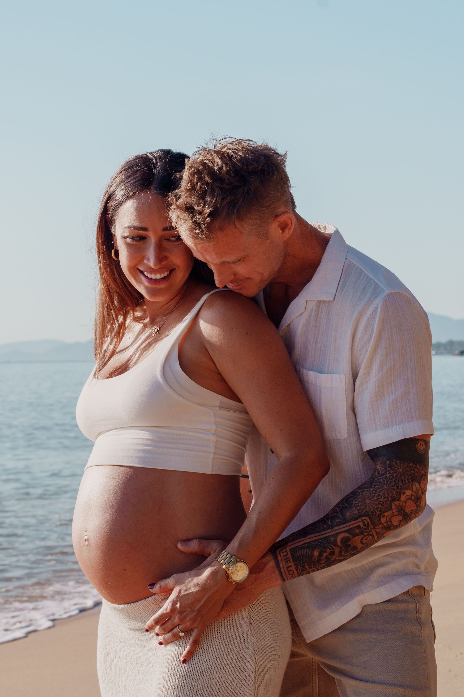 Tattooed man embracing smiling pregnant woman in white crop top on sunny beach by the ocean.