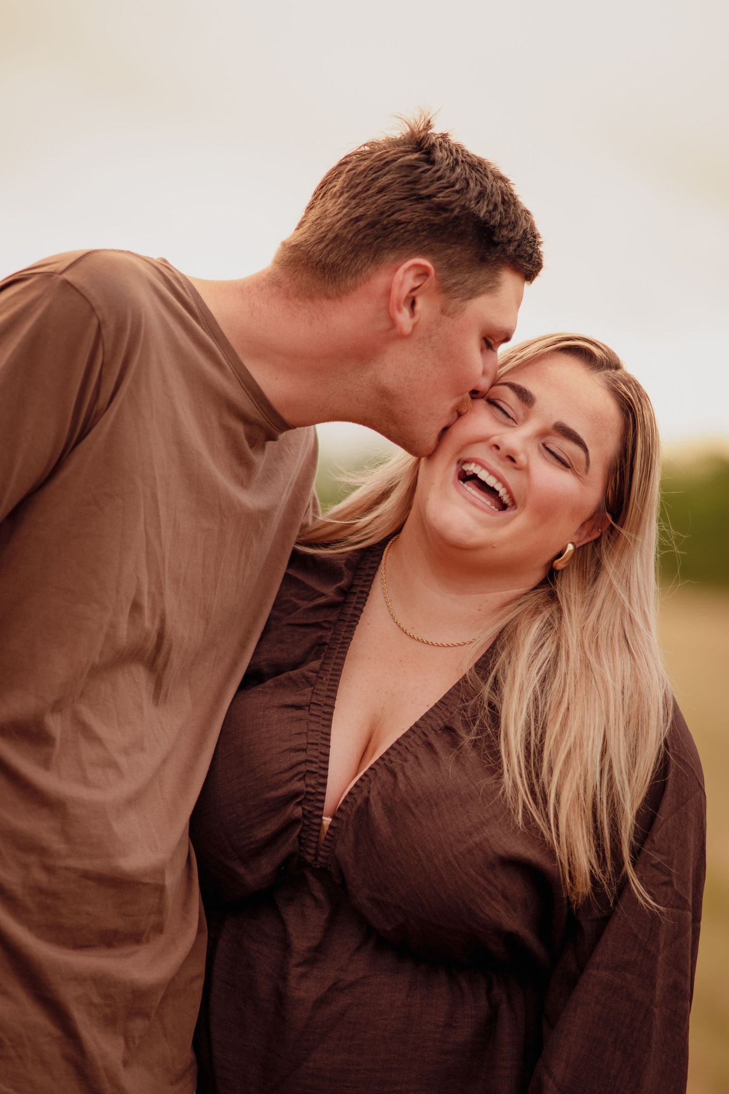 A man in a brown shirt kisses a laughing blonde woman on the cheek outdoors in a warm, romantic moment.