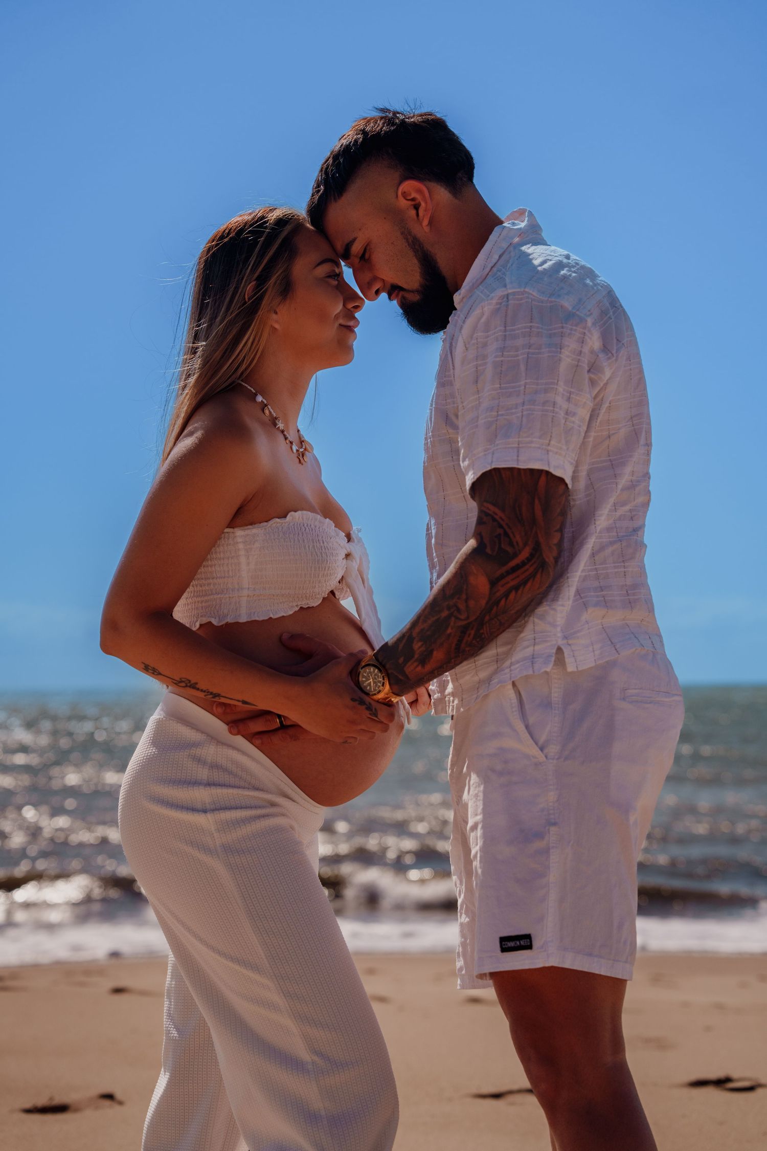 Expecting couple in white outfits tenderly touching pregnant belly on sunny beach with blue sky.