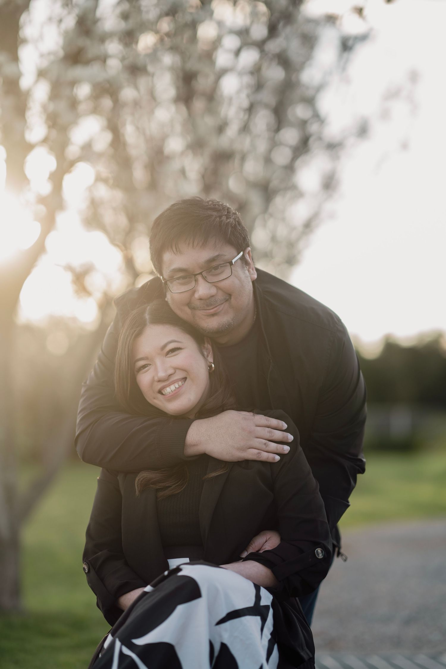 Smiling couple embracing warmly in a sunlit park with bokeh background.