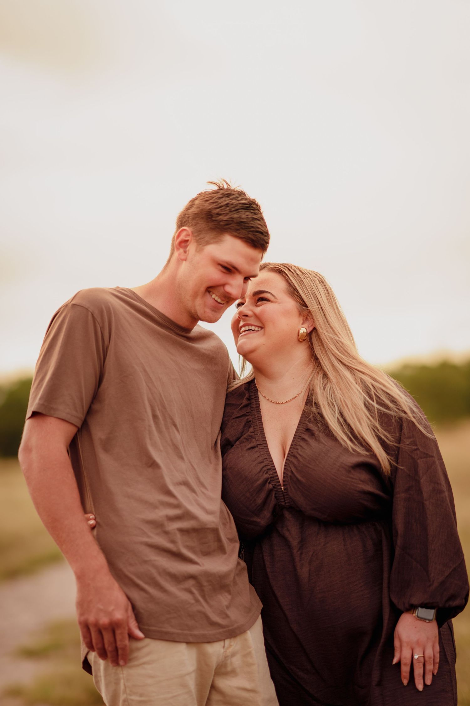 A smiling couple touches foreheads outdoors, the woman visibly pregnant in a dark dress, the man in a brown tee.