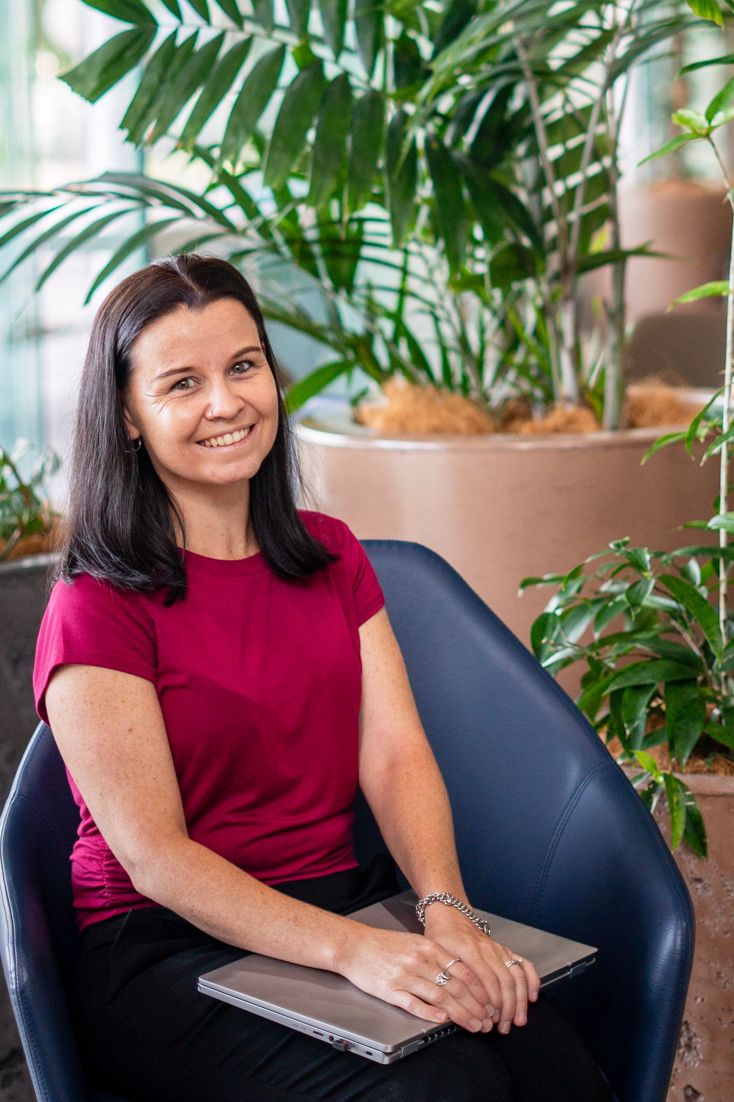 Smiling woman in red top sitting in blue chair with laptop, surrounded by lush green tropical plants indoors.