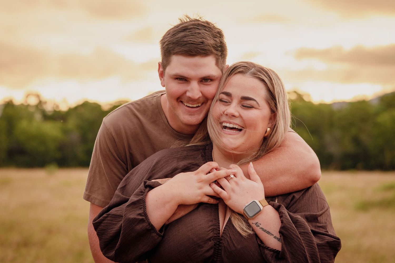 Couple laughing together in golden field at sunset, man hugging woman from behind in warm outdoor portrait.