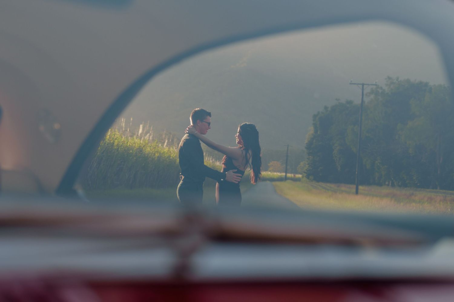 Romantic couple embraces on rural road viewed through vintage car window in soft hazy light.