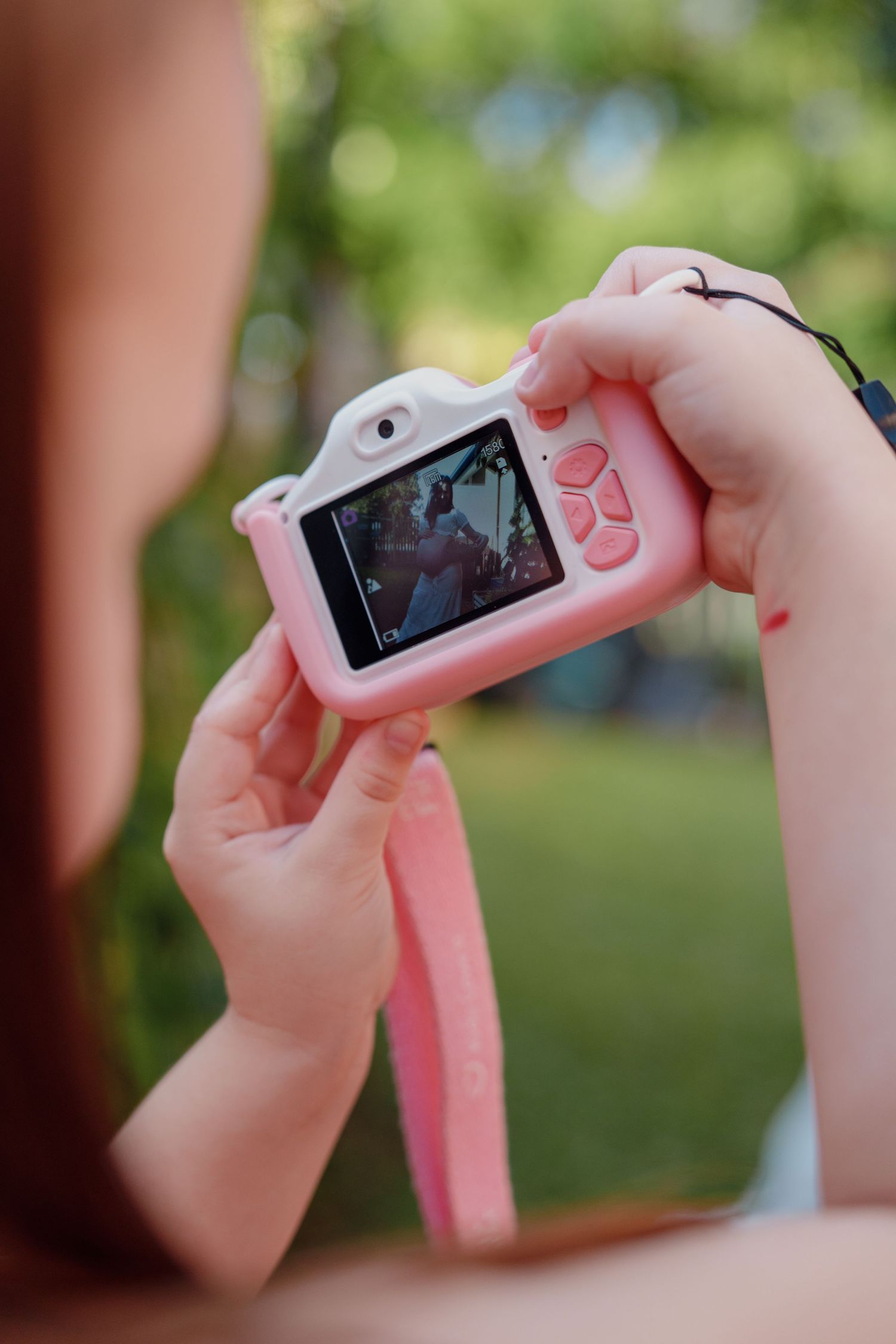 Child holds a pink toy camera outdoors, capturing photos on its small screen in a lush green garden setting.