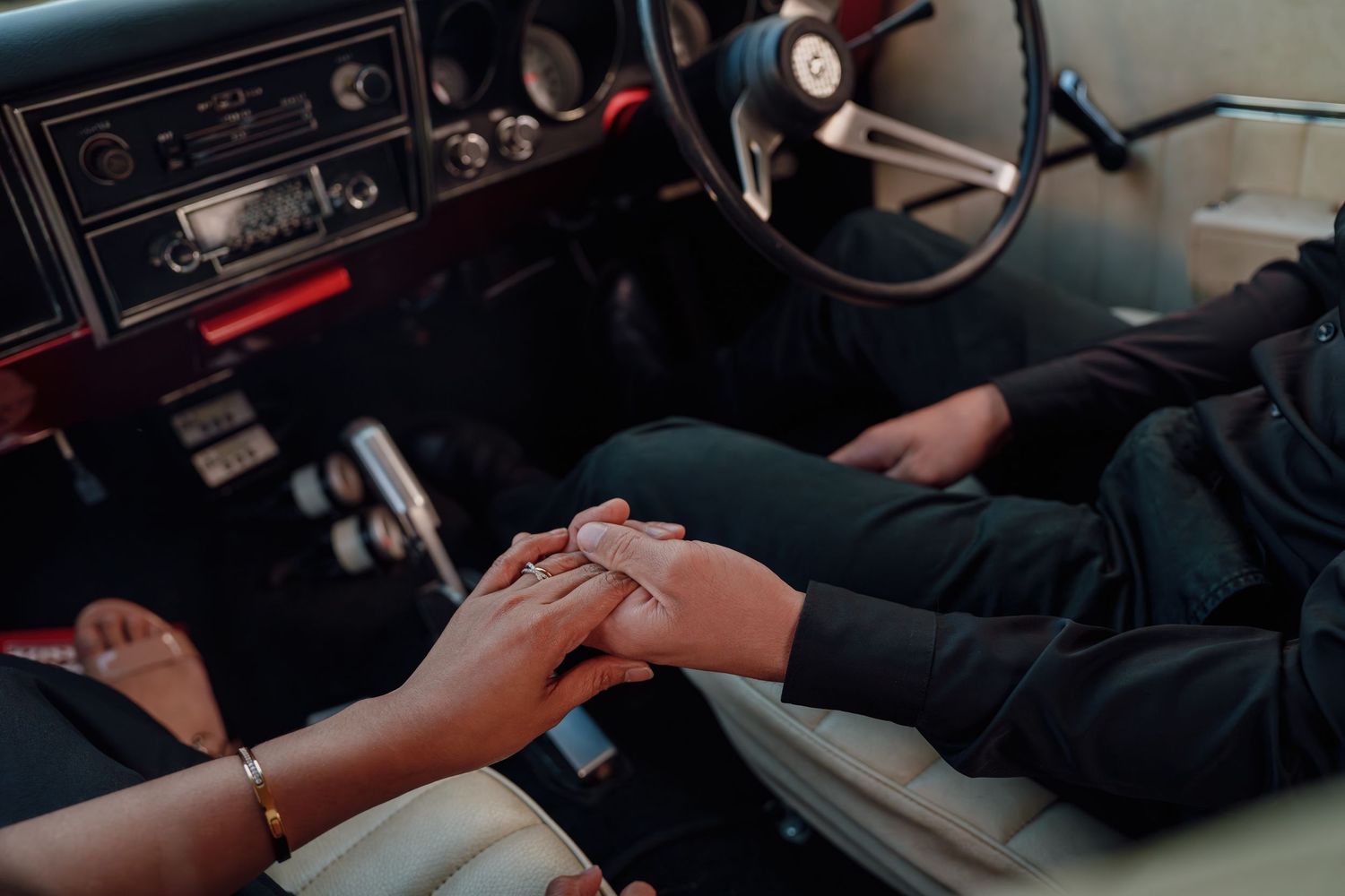 Couple holds hands tenderly inside vintage classic car interior with retro dashboard visible.