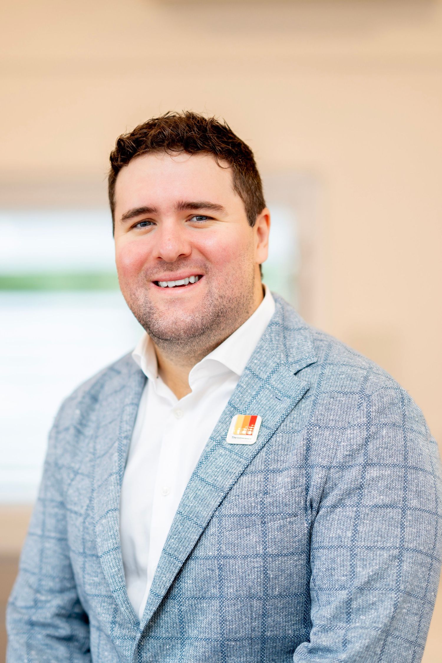 Smiling man in light blue plaid blazer with colorful lapel pin poses for professional headshot photo.