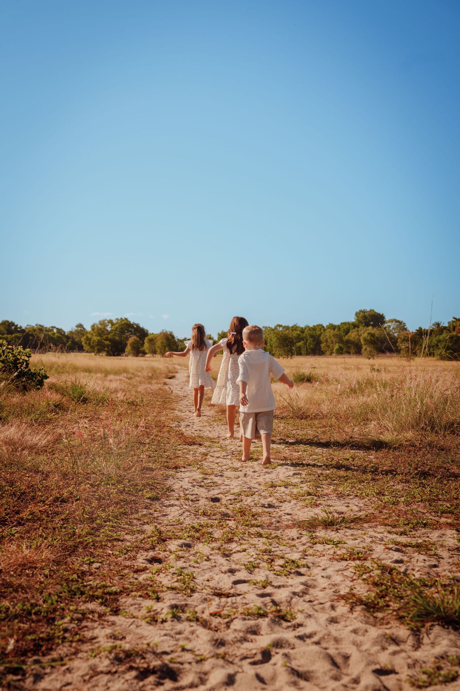 Four children walking barefoot along a sandy path through a golden field under a clear blue sky.