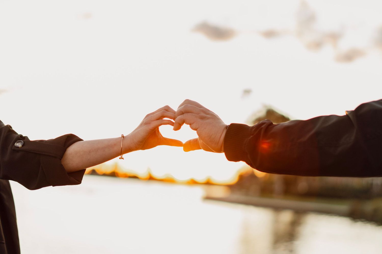 A couple forms a heart shape with their hands against a glowing golden sunset sky.