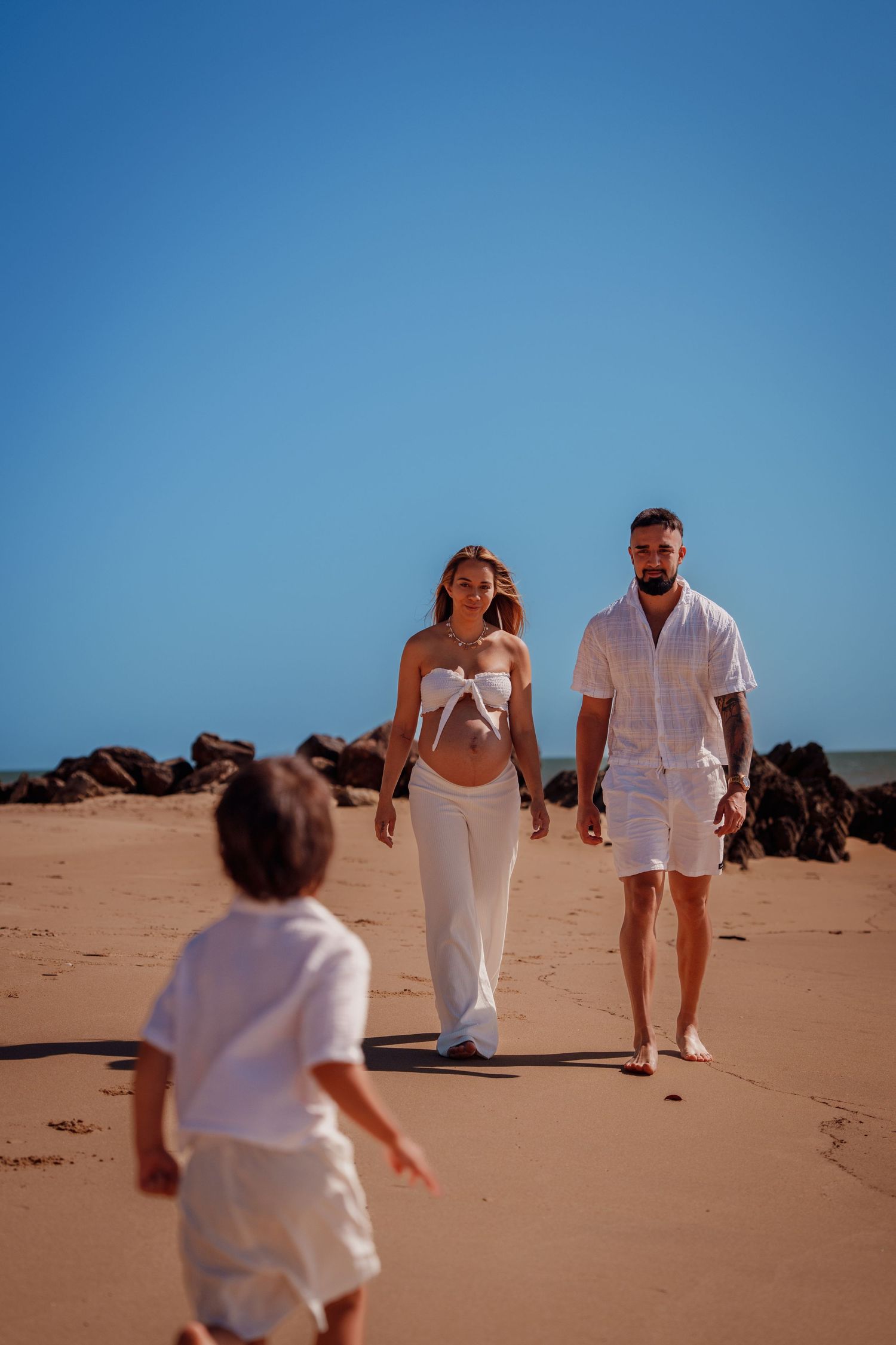 A family in white outfits walks on a sunny sandy beach, with a child running ahead and rocky cliffs in the background.