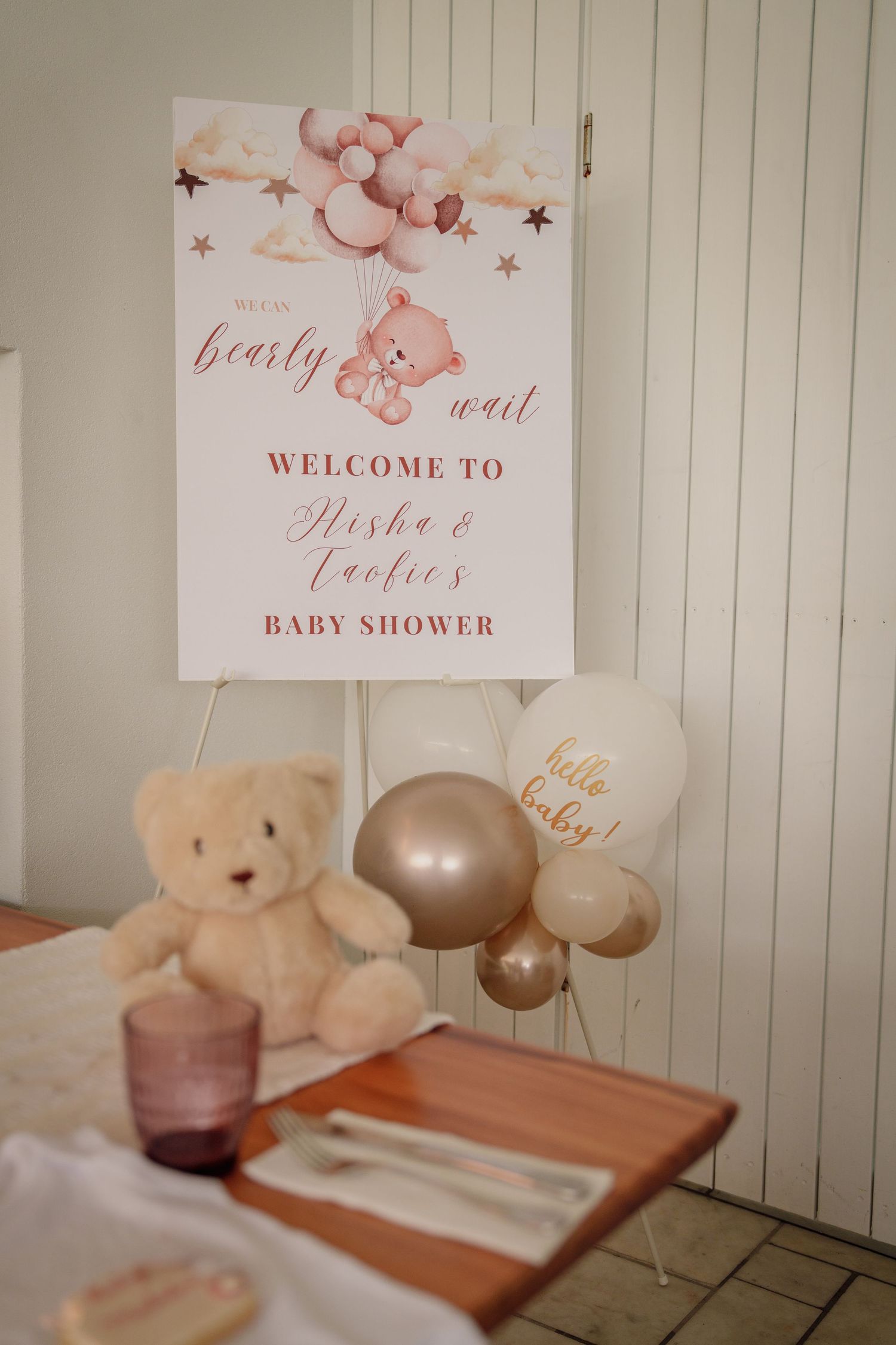 Baby shower welcome sign with teddy bear theme, gold and pink balloons, and plush bear centerpiece on decorated table.